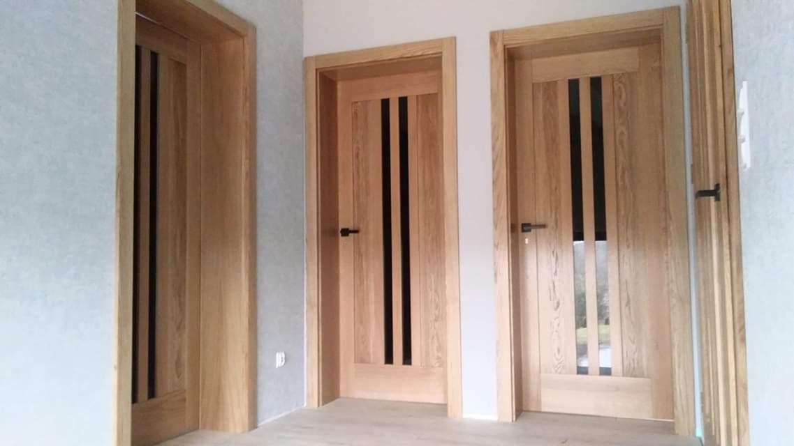 Trzy drewniane drzwi wewnętrzne z czarnymi, pionowymi wstawkami, zamontowane w jasnym pomieszczeniu z szarą ścianą.