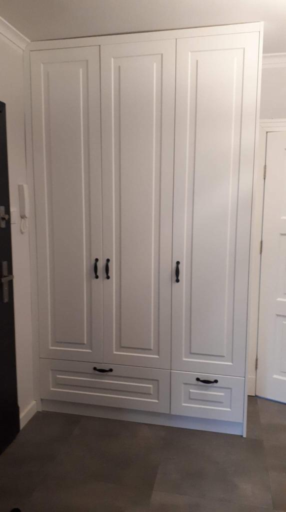 Trzydrzwiowa, biała szafa z czarnymi uchwytami, z dwiema szufladami w dolnej części, ustawiona w przedpokoju z szarą podłogą i białymi drzwiami w tle.