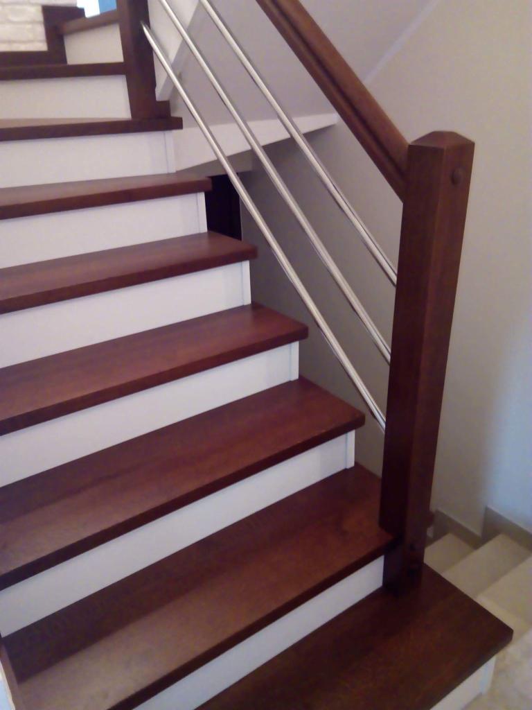 Nowoczesne schody z drewnianymi stopniami w odcieniu ciemnego brązu, białymi podstopnicami i balustradą ze stali nierdzewnej z drewnianym pochwytem.