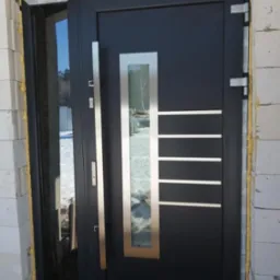 Ciemne drzwi wejściowe z długim, pionowym uchwytem i prostokątnym przeszkleniem z metalową ramką, zamontowane w surowym otworze murarskim z widoczną pianką uszczelniającą.