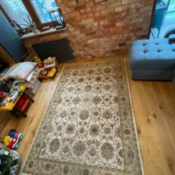 Wzorzysty dywan w odcieniach beżu i brązu leży na drewnianej podłodze w pokoju z ceglaną ścianą i niebieską sofą w tle, otoczony zabawkami dziecięcymi.