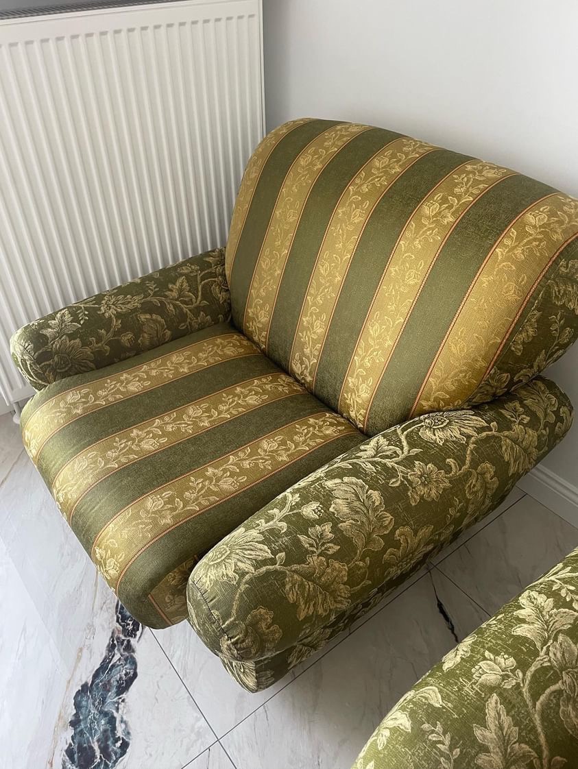 Zielono-złoty fotel tapicerowany w paski i kwiaty, stojący na marmurowej podłodze przy białym kaloryferze.