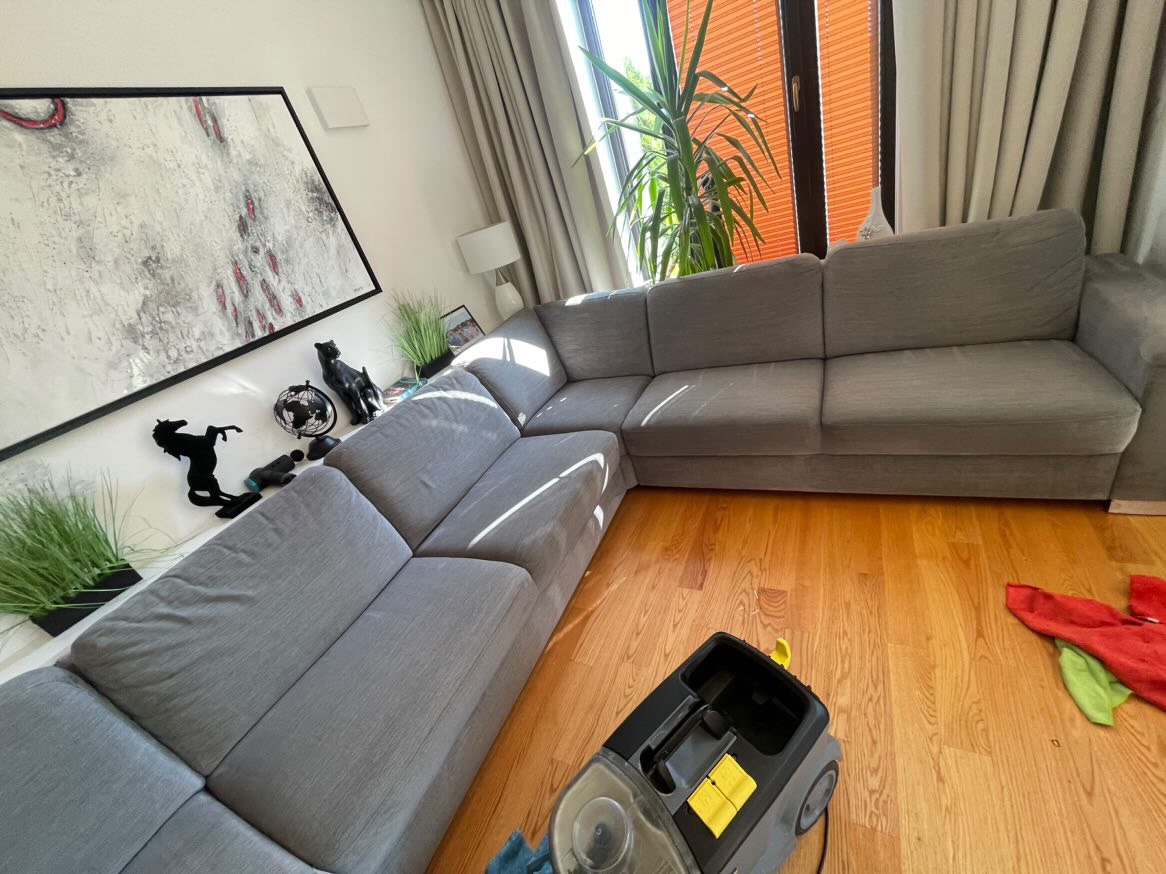 Szara, modułowa kanapa narożna w jasnym salonie w Warszawie, częściowo oświetlona słońcem. Na podłodze widoczny odkurzacz piorący, sugerujący proces czyszczenia. W tle okno z pomarańczowymi...