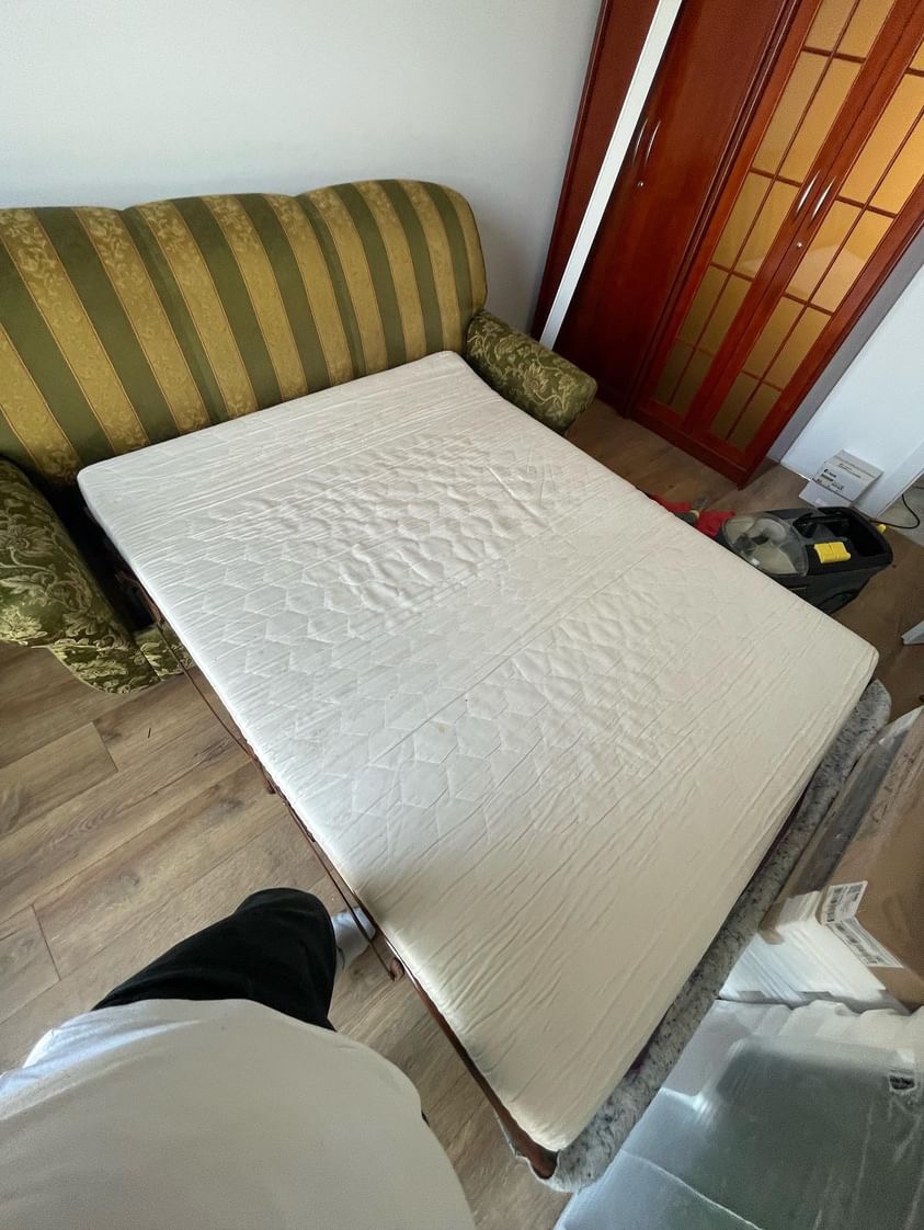 Rozłożona kanapa z materacem, widoczne wzorzyste obicie kanapy w odcieniach zieleni i beżu oraz biały materac z delikatnym wzorem, na podłodze leży odkurzacz.