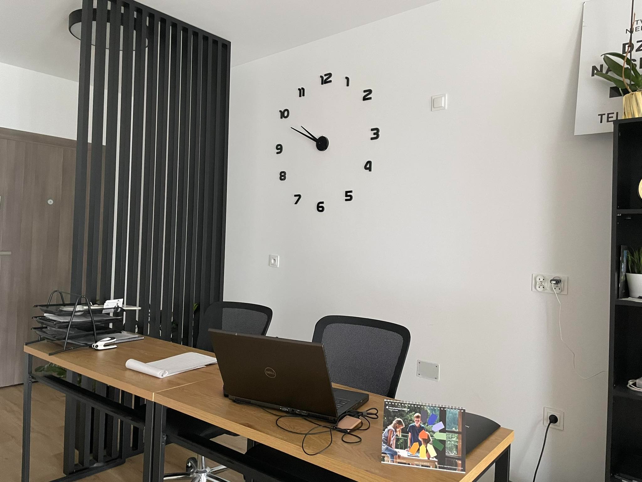 Nowoczesne biuro z drewnianym biurkiem, czarnym laptopem Dell, kalendarzem i minimalistycznym zegarem ściennym z dużymi, czarnymi cyframi.