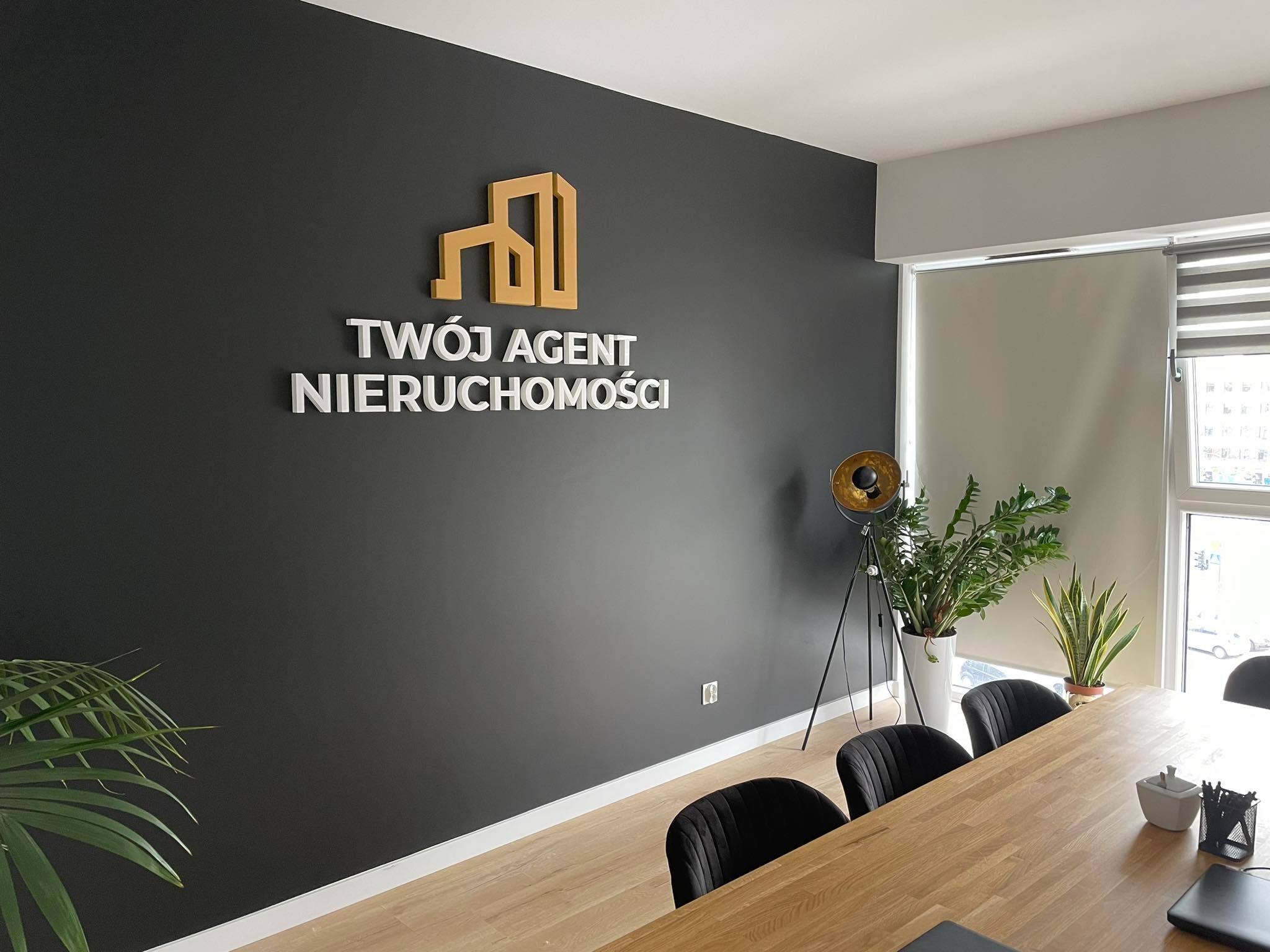 Wnętrze biura agenta nieruchomości z logo firmy 'Twój Agent Nieruchomości' na ciemnej ścianie, oświetlone złotą lampą stojącą i z roślinami doniczkowymi w tle.