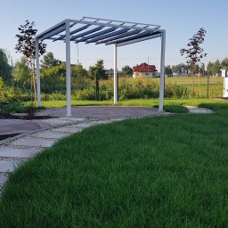 Green Media - architektura krajobrazu - Nowoczesna pergola z jasną konstrukcją w zadbanym ogrodzie z trawnikiem, ścieżką z kamieni i ozdobnymi nasadzeniami. W tle widoczny dom jednorodzinny.