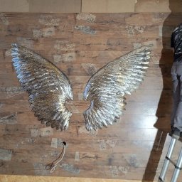 KostuchArt Studio - Instalacja dekoracyjnych, srebrnych skrzydeł na ścianie z drewnianymi panelami z nadrukiem tablic rejestracyjnych, wykonywana przez mężczyznę na drabinie.