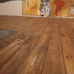 KostuchArt Studio - Podłoga z desek w odcieniach brązu, z widocznym rysunkiem słojów drewna, na ścianie malowidło przedstawiające samolot i motocyklistę na tle mapy świata.