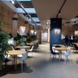 KostuchArt Studio - Jasne wnętrze kawiarni lub biura z drewnianymi panelami, okrągłymi stolikami i nowoczesnymi krzesłami. Oświetlenie punktowe na suficie, rośliny doniczkowe w rogu kadru. Widoczne osoby w oddali.