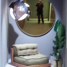 KostuchArt Studio - Minimalistyczny pokój z beżowym fotelem, okrągłym lustrem odbijającym postać i rośliną doniczkową, oświetlony nowoczesną lampą wiszącą.