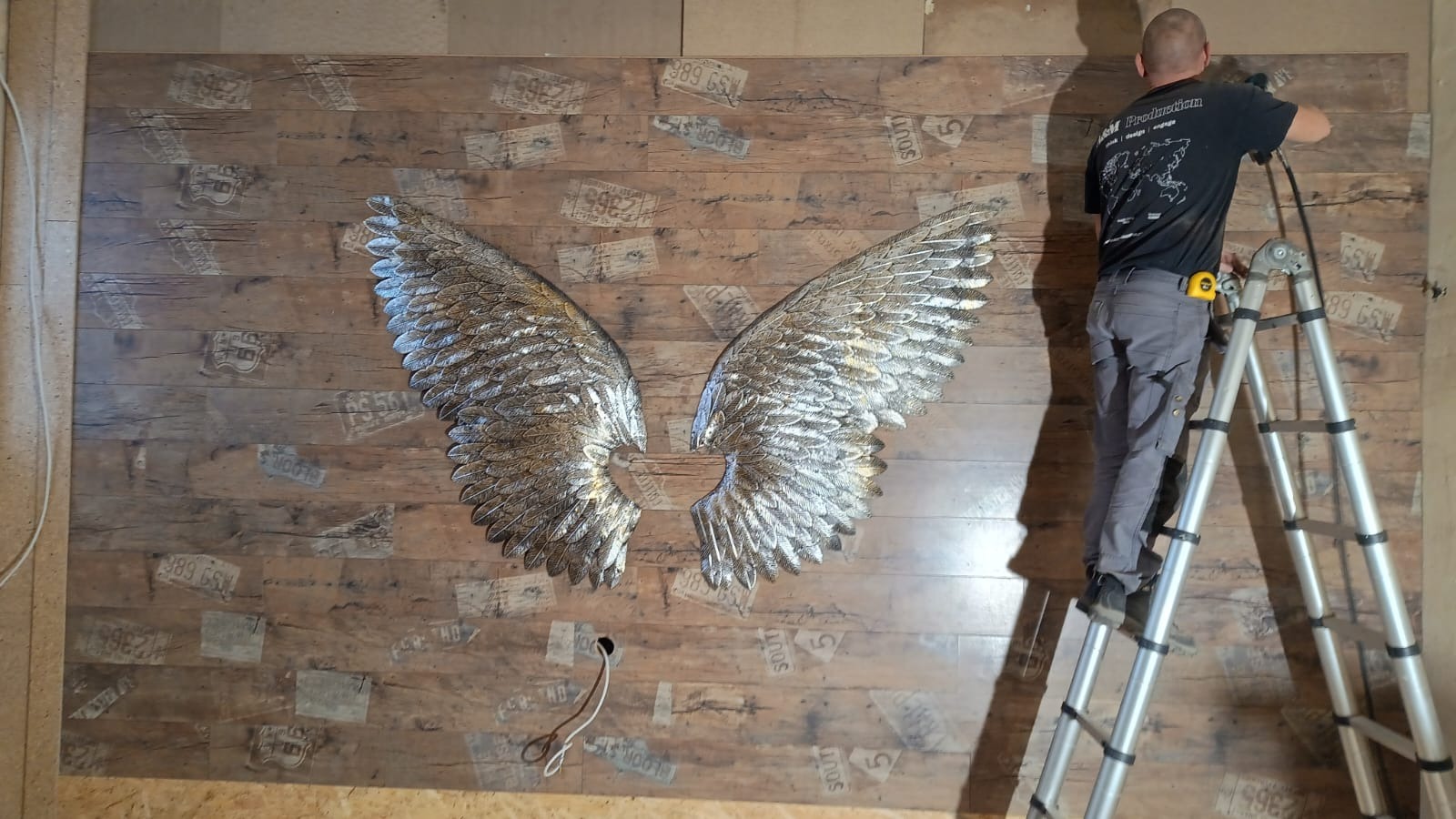 Instalacja dekoracyjnych, srebrnych skrzydeł na ścianie z drewnianymi panelami z nadrukiem tablic rejestracyjnych, wykonywana przez mężczyznę na drabinie.
