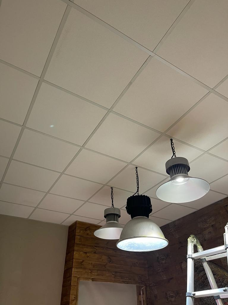 Widok w górę na sufit kasetonowy z trzema przemysłowymi lampami wiszącymi na łańcuchach, drewniane panele ścienne w tle i fragment drabiny malarskiej.