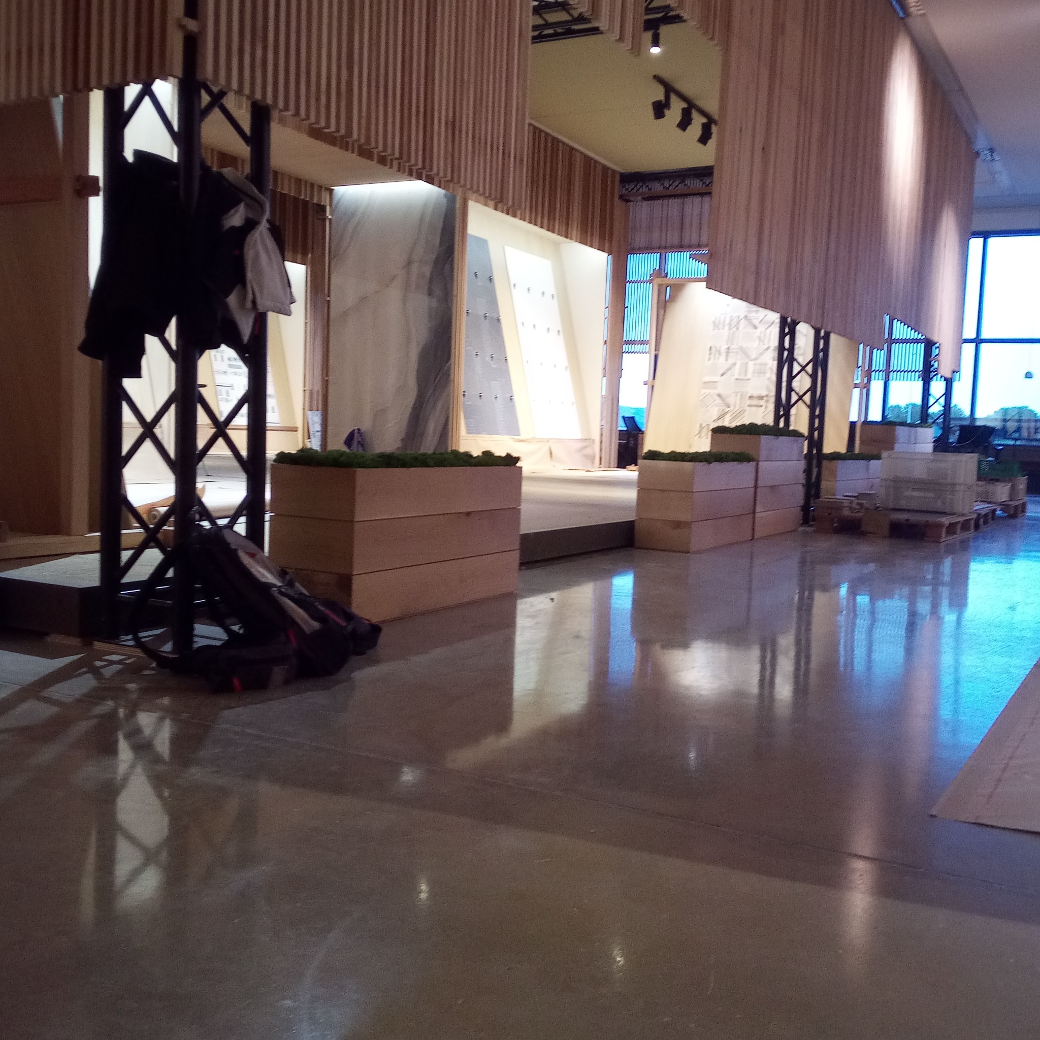 Wnętrze przestrzeni wystawienniczej z panelami dekoracyjnymi, drewnianymi elementami konstrukcyjnymi i donicami z zielenią, widoczne ślady po pracach wykończeniowych, torba robocza i ubrania...