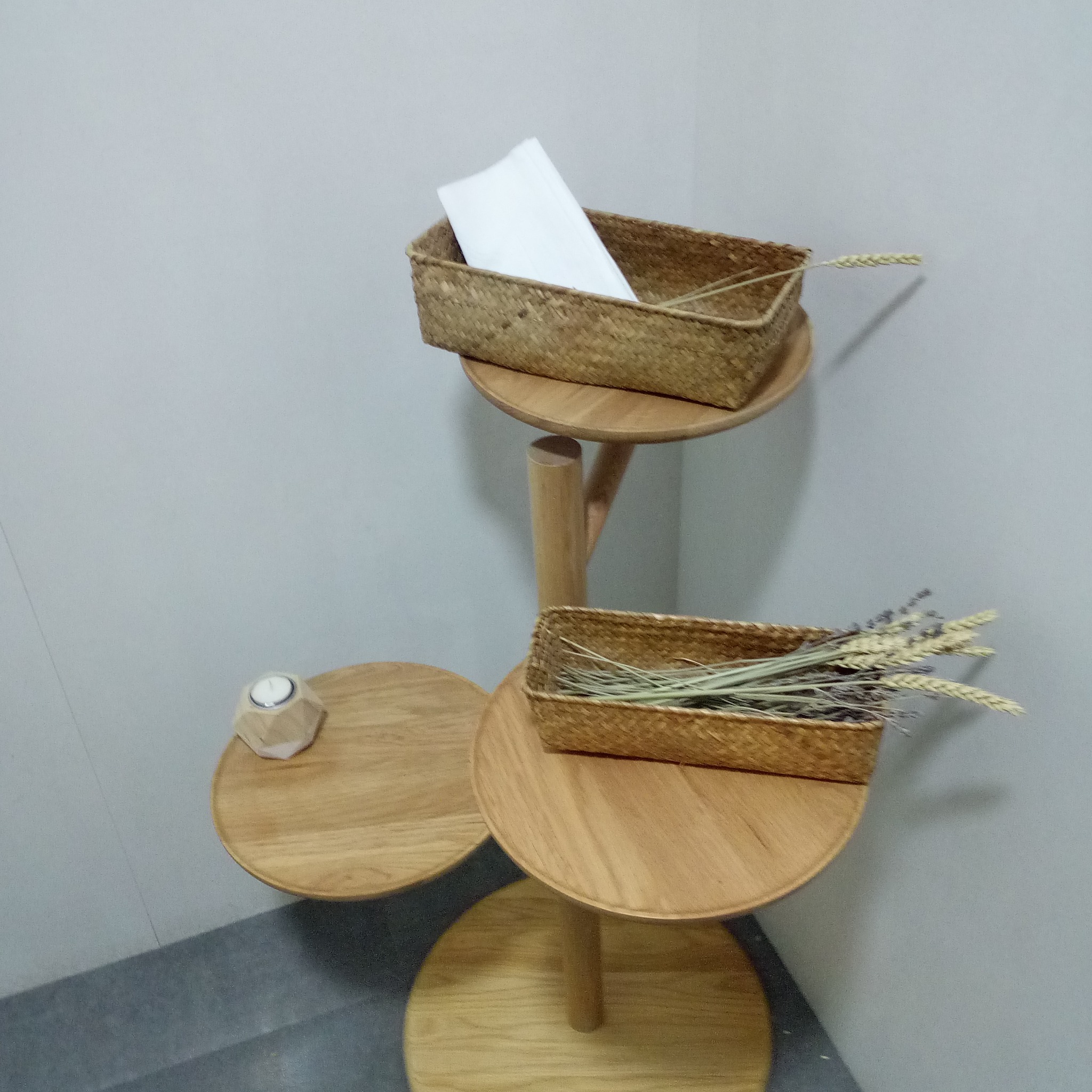 Drewniany, trójpoziomowy stolik dekoracyjny z wiklinowymi koszykami, świecą i suszonymi kwiatami w rogu pomieszczenia z szarą ścianą.