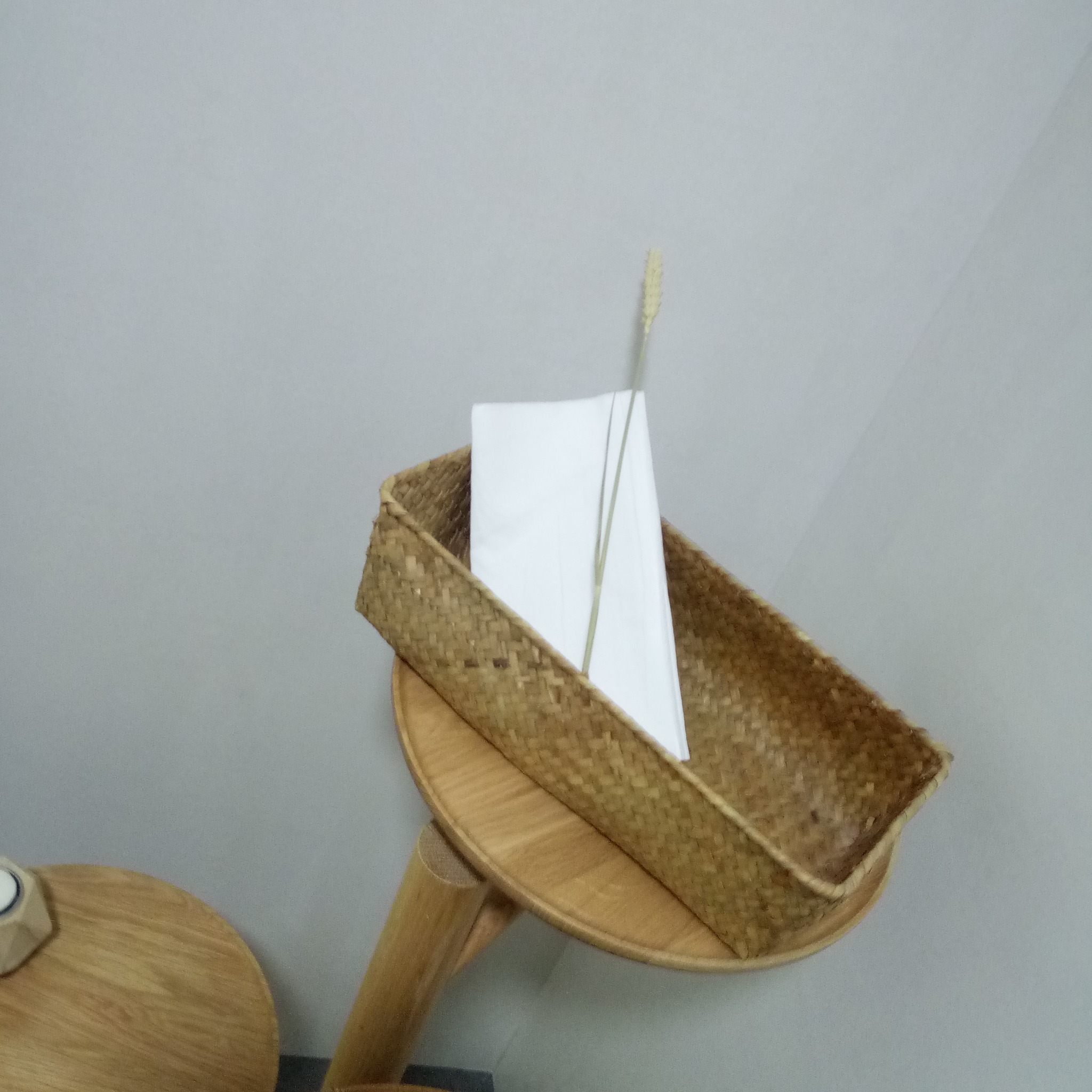 Dekoracyjny koszyk z trawy morskiej na drewnianym stoliku, zawierający białe tekstylia i źdźbło trawy, na tle szarej ściany.