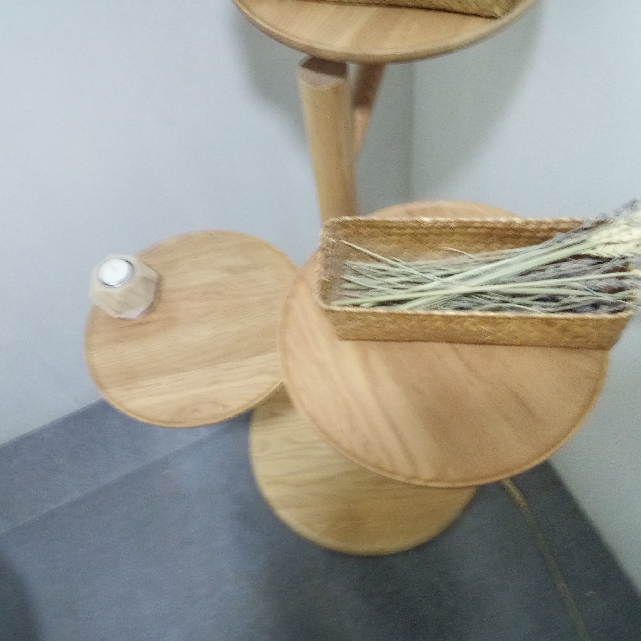 Trzypoziomowy, drewniany stolik pomocniczy z okrągłymi blatami, na którym znajduje się świeca i wiklinowy koszyk z suchymi trawami, ustawiony na szarym dywanie.