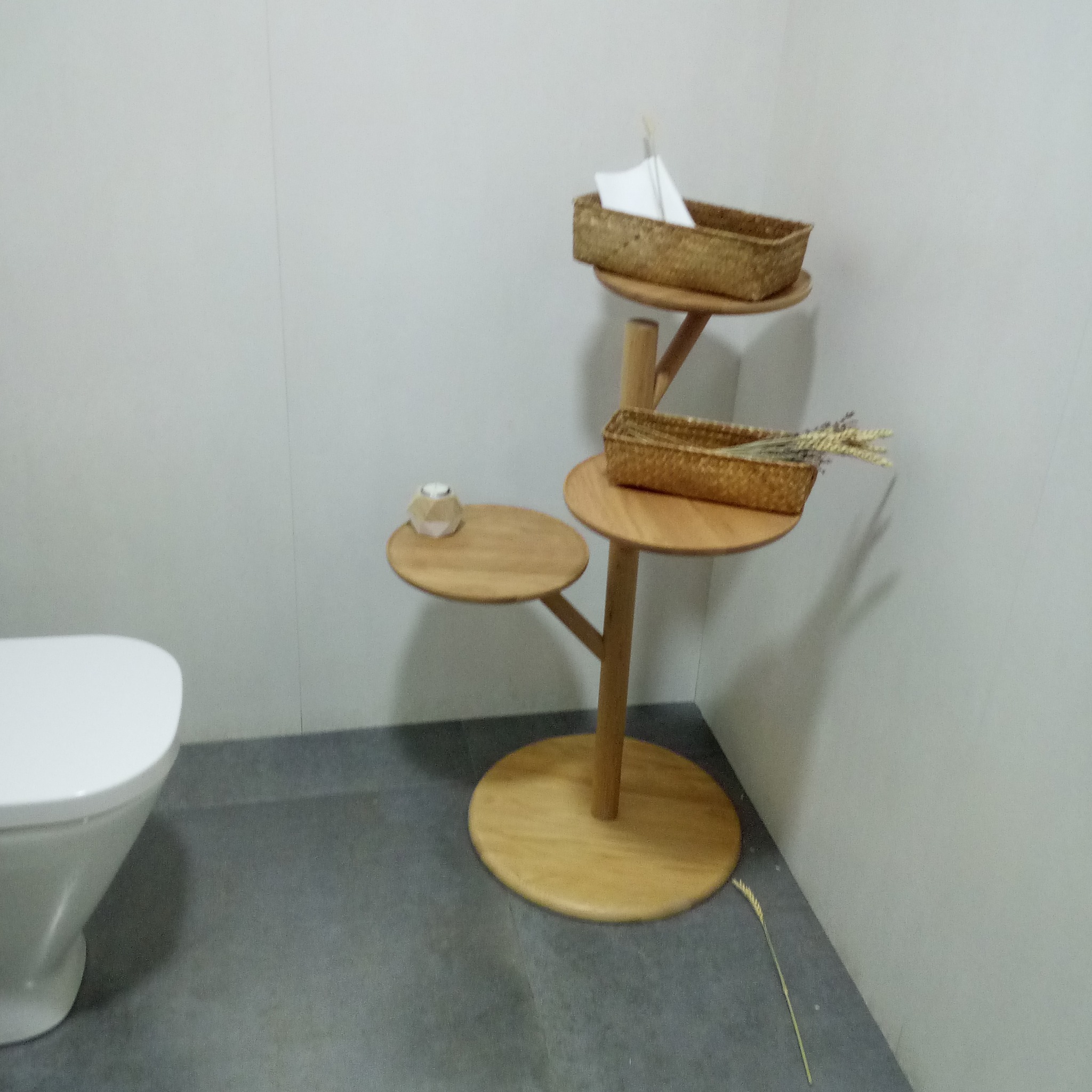 Minimalistyczna łazienka z drewnianym regałem na akcesoria, na którym znajdują się wiklinowe koszyki z papierem toaletowym i suszonymi kwiatami, obok toaleta.