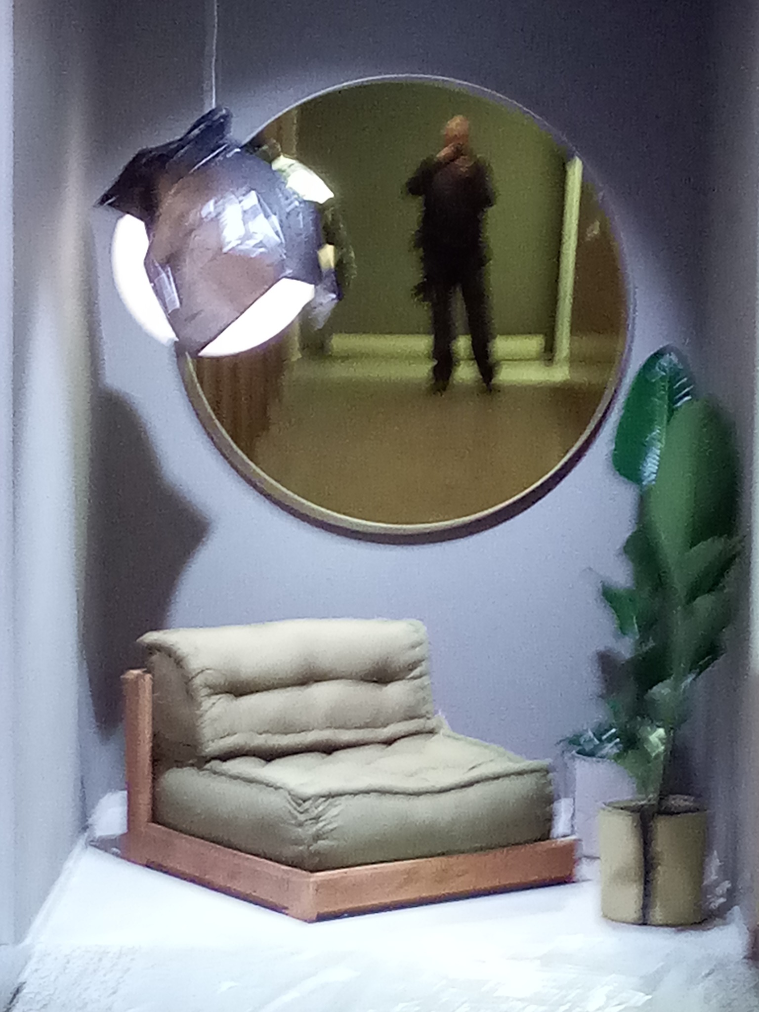 Minimalistyczny pokój z beżowym fotelem, okrągłym lustrem odbijającym postać i rośliną doniczkową, oświetlony nowoczesną lampą wiszącą.