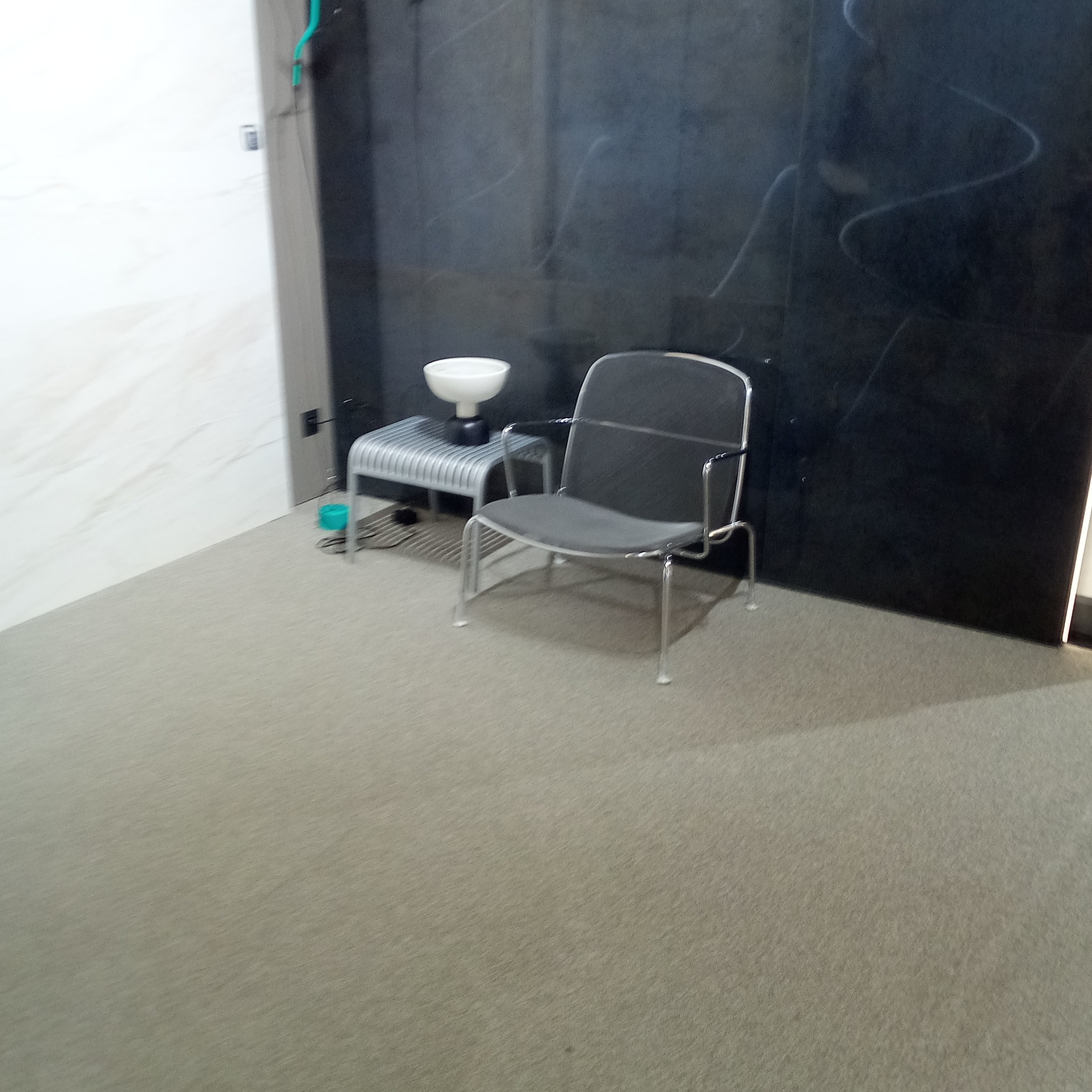 Wnętrze pokoju z ciemną, marmurkową ścianą, jasną podłogą, metalowym krzesłem i stolikiem z lampą.