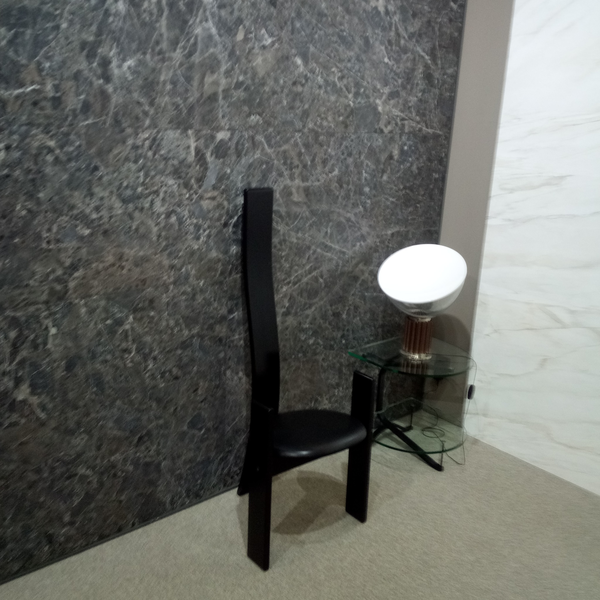 Fragment wykończonego wnętrza z ciemnoszarą marmurkową ścianą, czarnym krzesłem o wysokim oparciu i szklanym stolikiem z lampką.