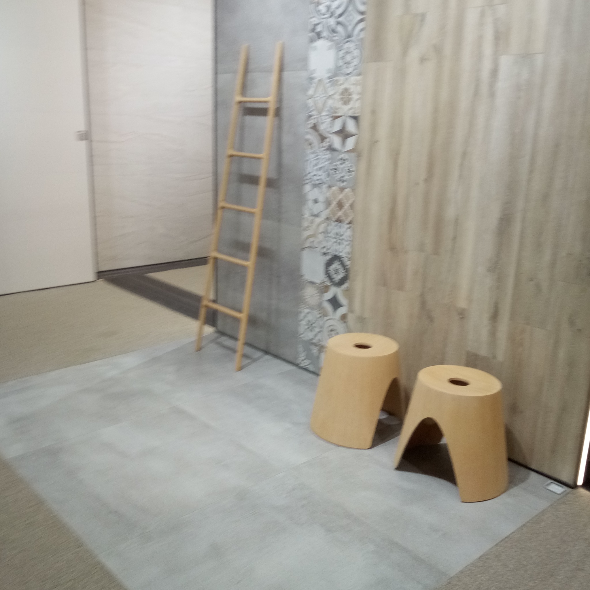 Wnętrze z drewnianą drabinką dekoracyjną, dwoma stołkami w kształcie walca z otworem, panelami ściennymi o różnej fakturze i wzorach oraz szarą podłogą.