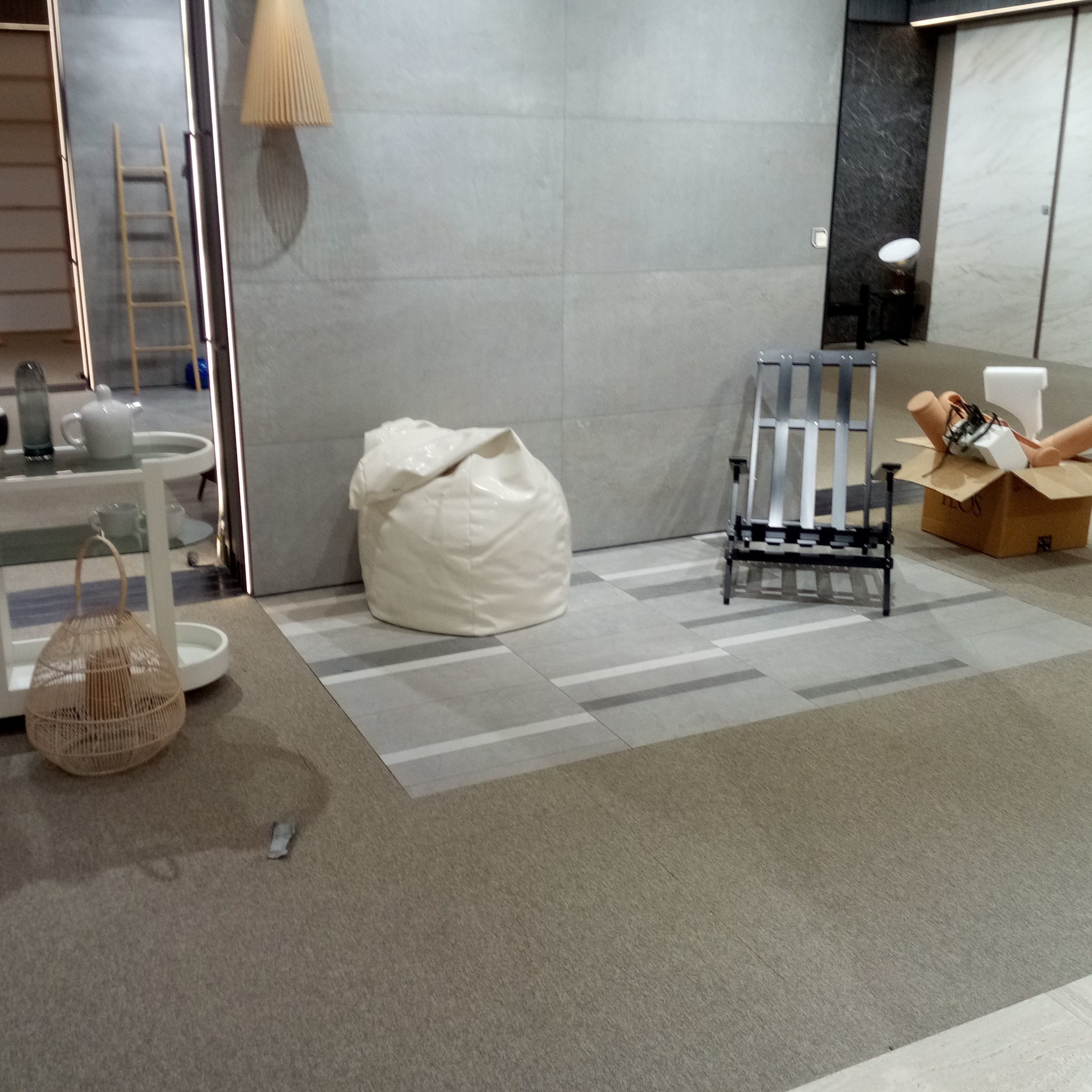 Wnętrze z szarymi płytkami podłogowymi z białymi paskami, białym pufem, metalowym krzesłem i kartonem z akcesoriami, elementami dekoracyjnymi i lampą wiszącą w tle.