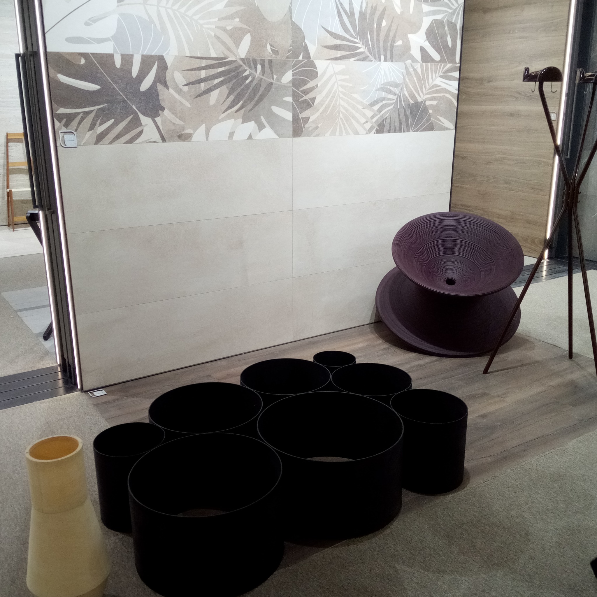 Ekspozycja płytek ceramicznych z motywem liści w neutralnych kolorach, uzupełniona o dekoracyjne elementy wystroju wnętrz, takie jak czarne cylindryczne formy i purpurowa, spiralna donica.