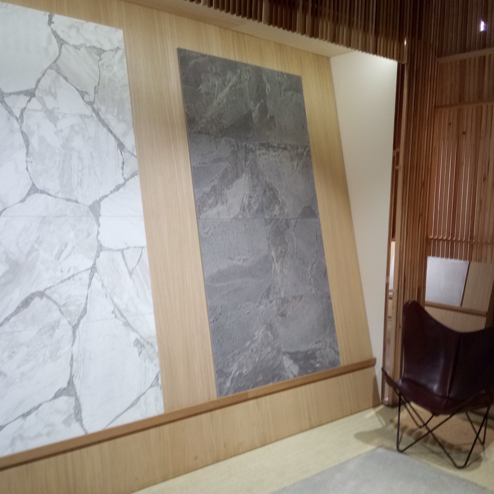 Ekspozycja dwóch paneli ściennych o różnych wzorach: biały z szarymi żyłkami imitujący marmur i szary imitujący kamień, umieszczonych na drewnianej ścianie w pomieszczeniu z krzesłem.