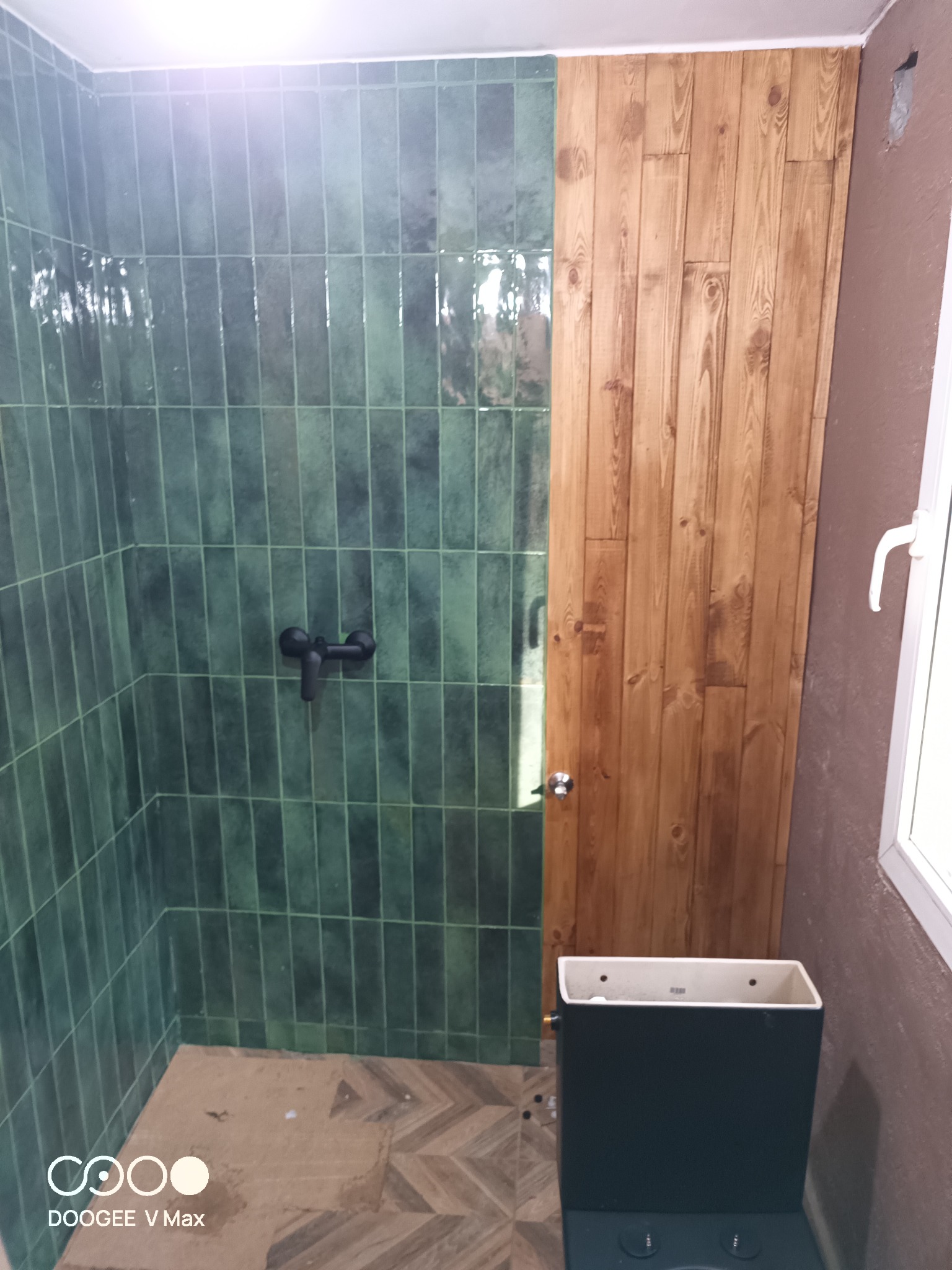 Wykończona łazienka z zielonymi, pionowymi płytkami w strefie prysznica, drewnianą boazerią na ścianie obok, czarną armaturą i widoczną, częściowo zamontowaną toaletą.