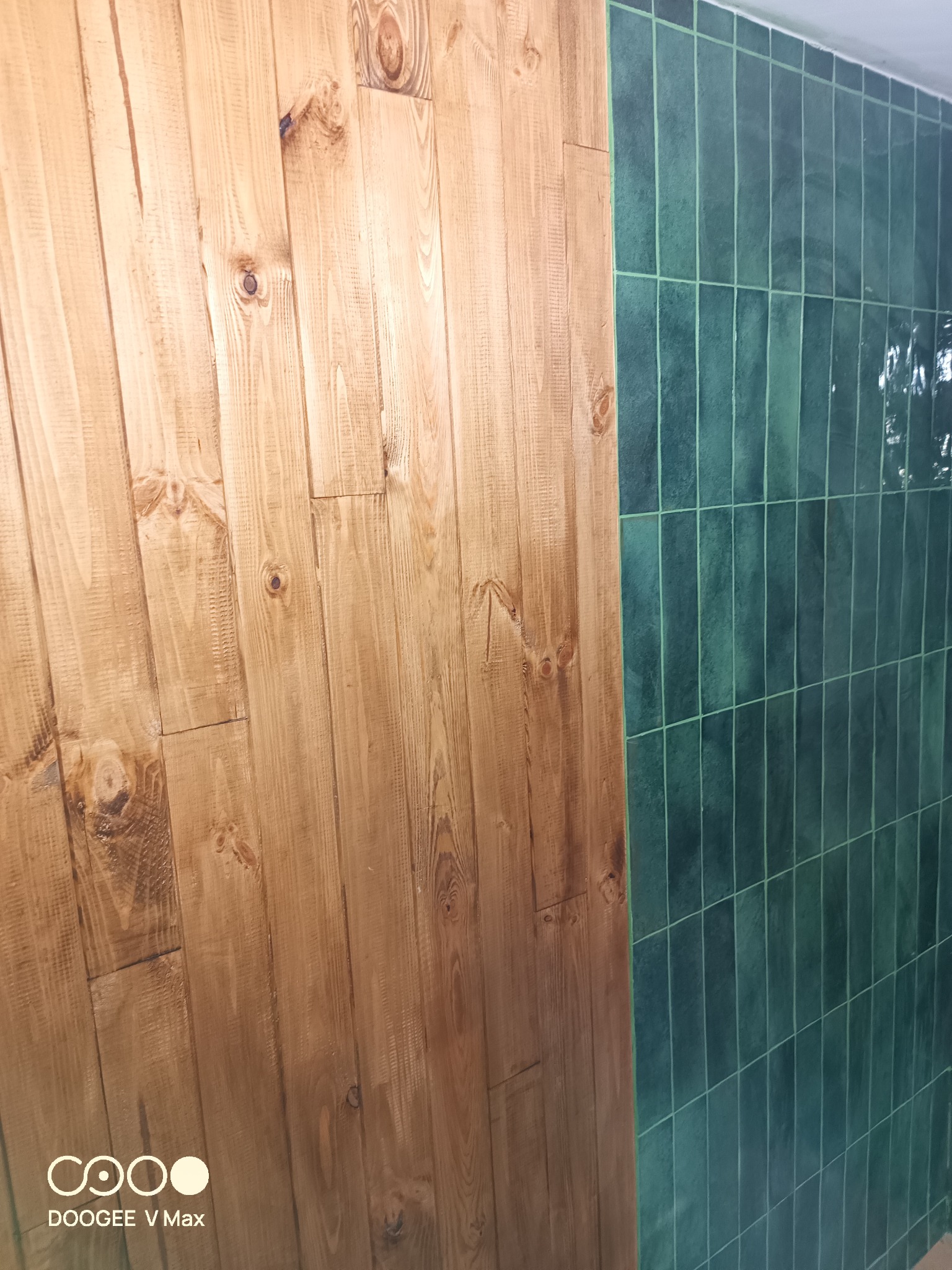 Fragment ściany z wykończeniem z pionowych desek z widocznymi słojami drewna, sąsiadującej z ścianą wyłożoną wąskimi, pionowymi, zielonymi płytkami.