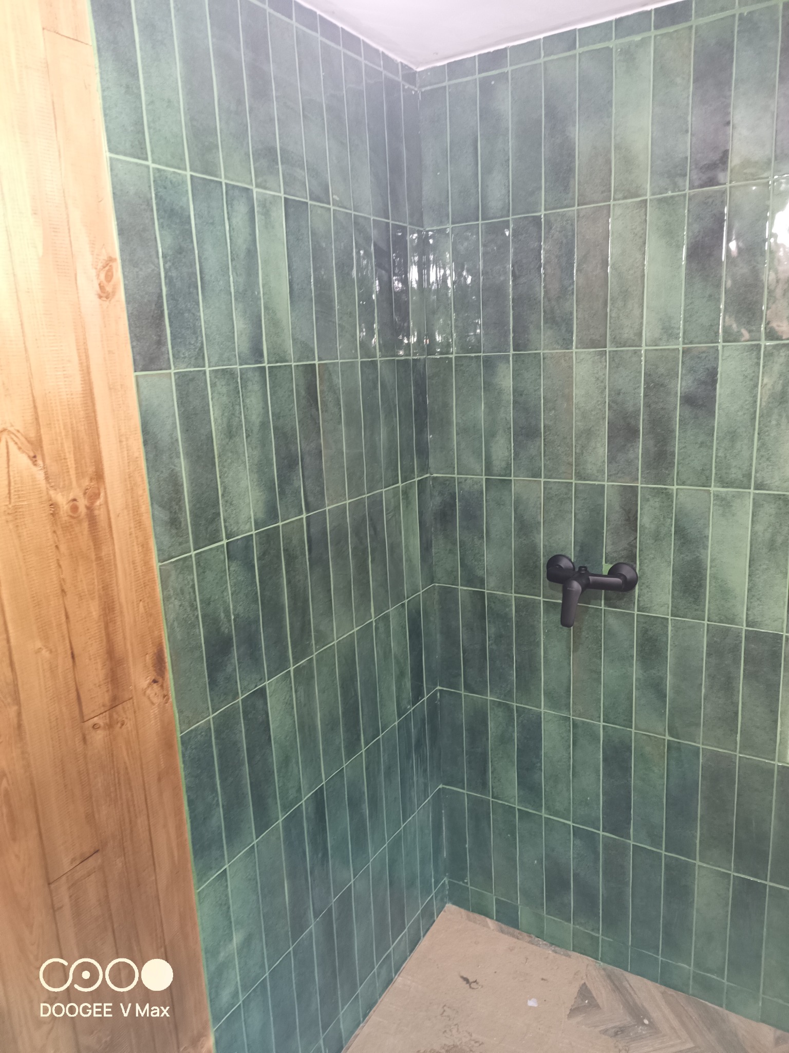 Ściana kabiny prysznicowej wyłożona pionowymi, zielonymi płytkami w odcieniu butelkowej zieleni z czarną baterią prysznicową i fragmentem drewnianej obudowy.