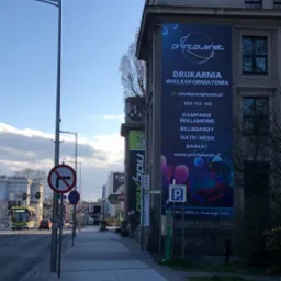 Billboard z reklamą drukarni wielkoformatowej Print Planet umieszczony na bocznej ścianie budynku wzdłuż ulicy, widoczne znaki drogowe i przejeżdżający autobus.