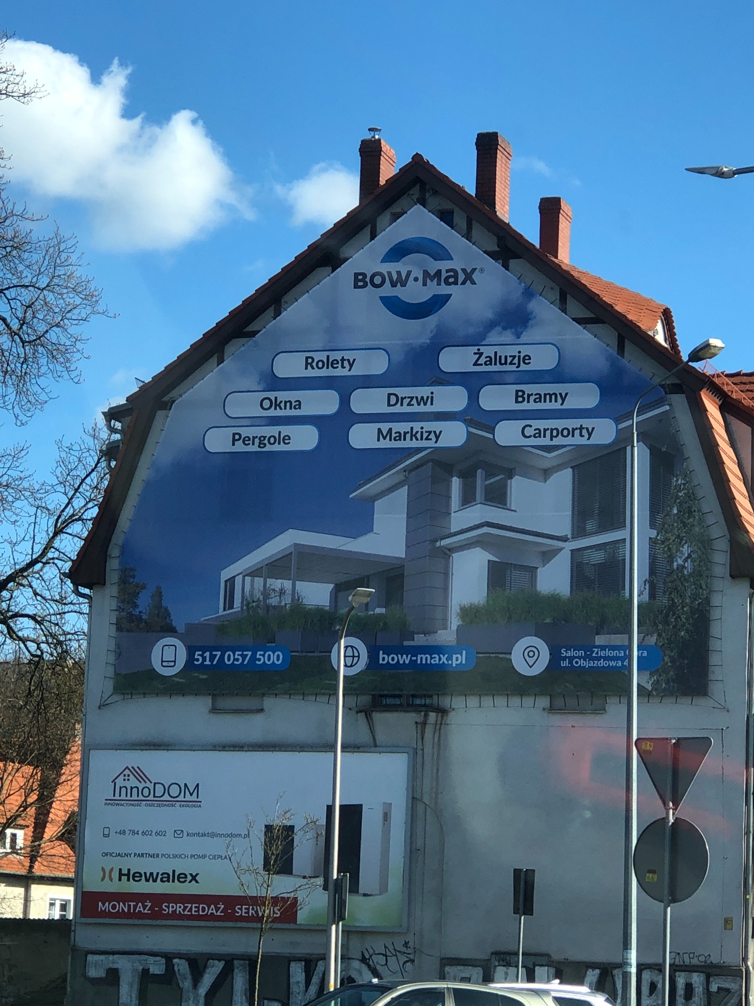 Reklama wielkoformatowa na ścianie budynku z ofertą rolet, żaluzji, okien, drzwi, bram, pergoli, markiz i carportów firmy Bow-Max oraz logiem InnoDOM i Hewalex.