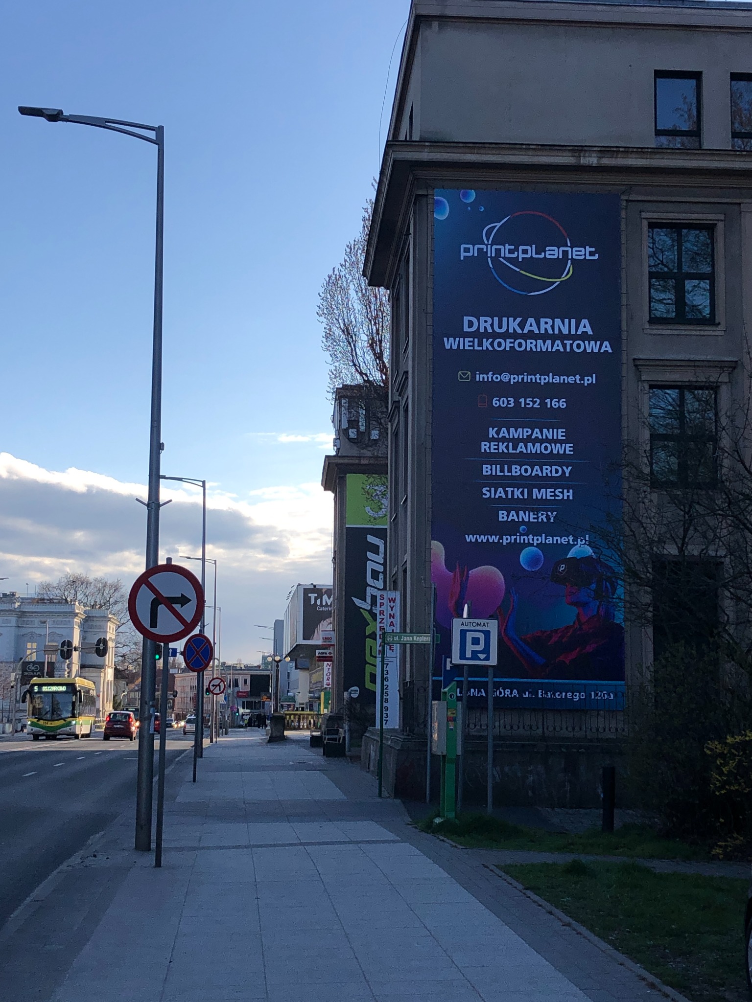 Billboard z reklamą drukarni wielkoformatowej Print Planet umieszczony na bocznej ścianie budynku wzdłuż ulicy, widoczne znaki drogowe i przejeżdżający autobus.