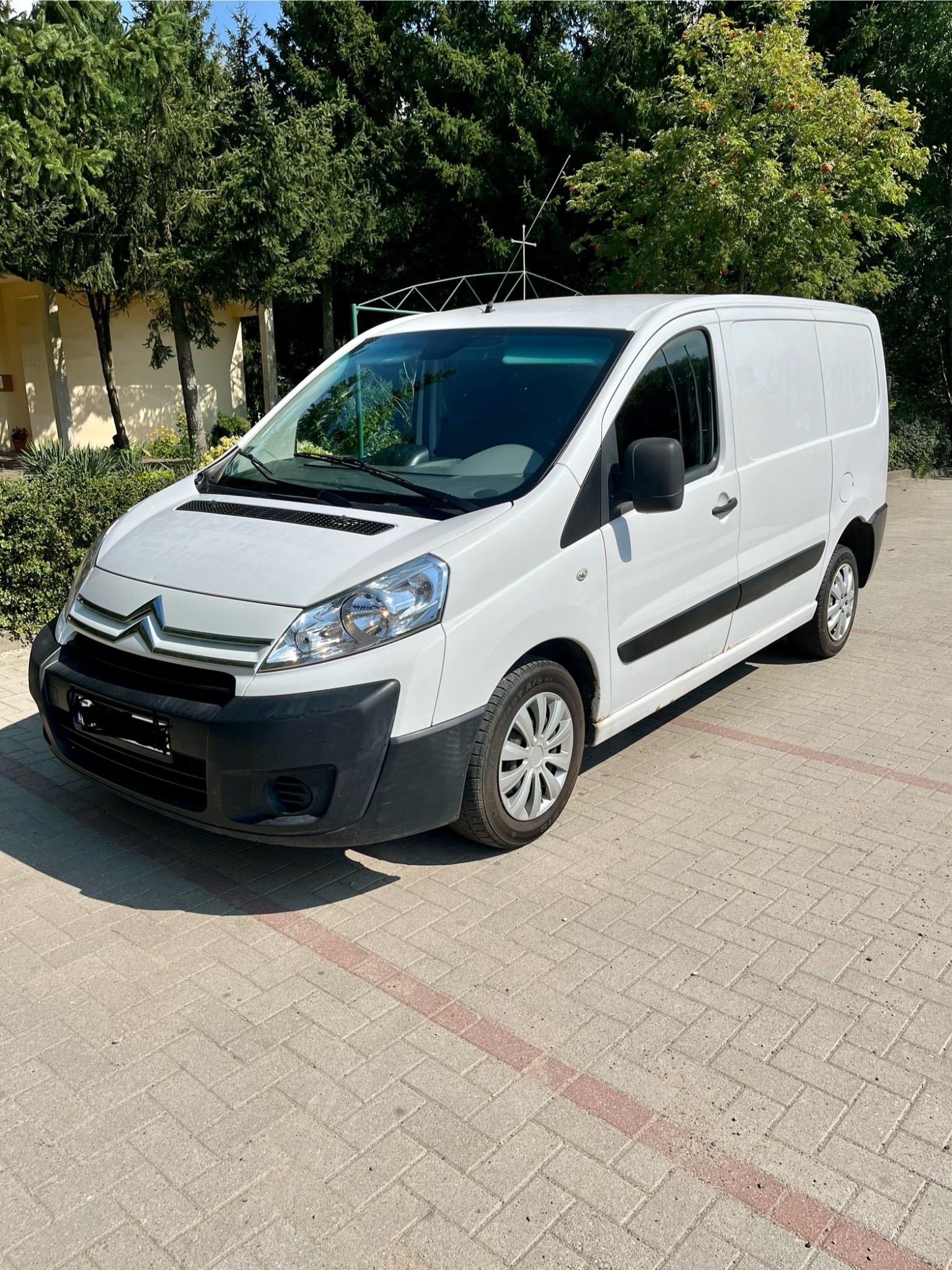 Biały samochód dostawczy Citroen Jumpy zaparkowany na brukowanym placu z widocznymi drzewami w tle.