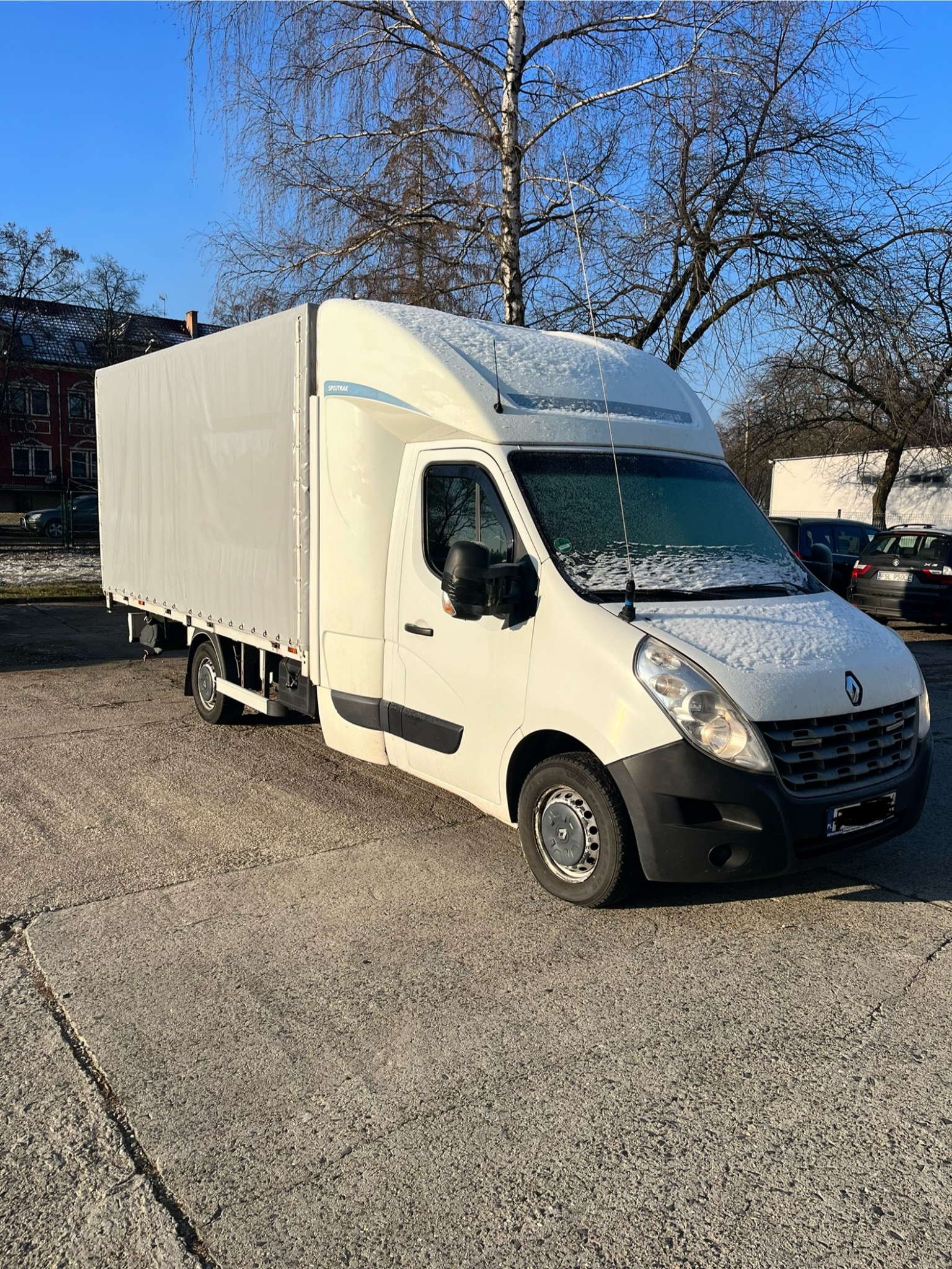 Biały samochód dostawczy Renault Master z plandeką, lekko oszroniony, zaparkowany na betonowym placu w zimowy dzień, z drzewami bez liści w tle.