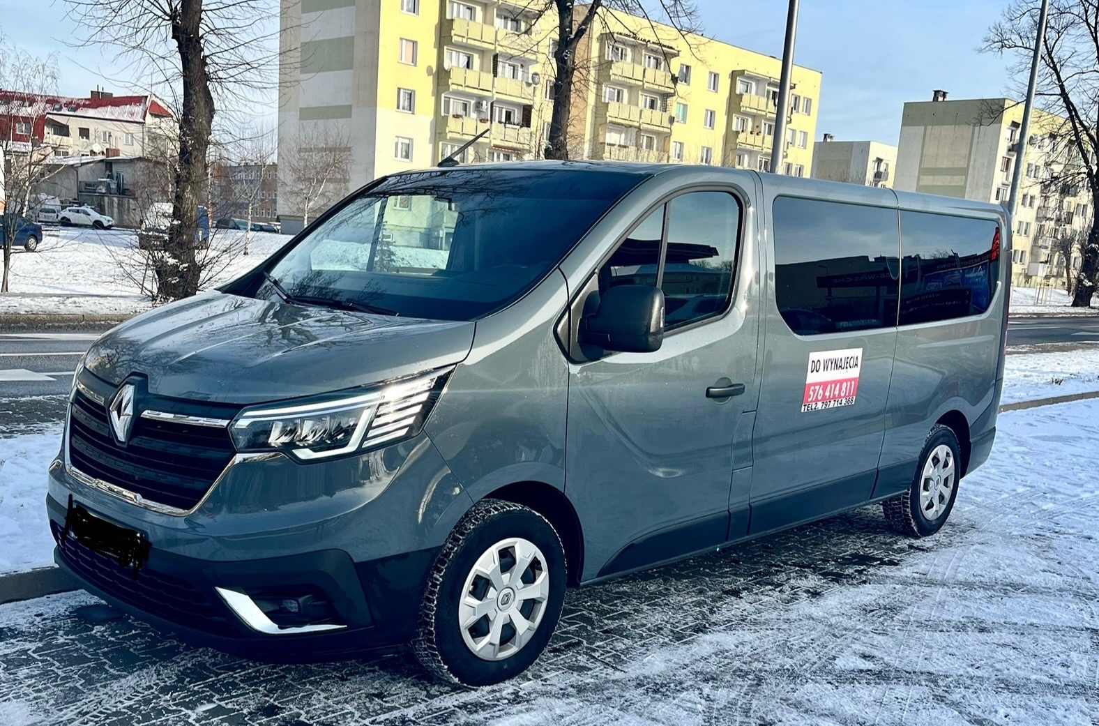 Szary Renault Trafic z naklejką 'Do wynajęcia' na tle miejskiego krajobrazu w zimowej scenerii.