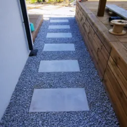 Ścieżka z betonowych płyt ułożona na szarym żwirze, prowadząca wzdłuż ściany budynku i drewnianego tarasu.