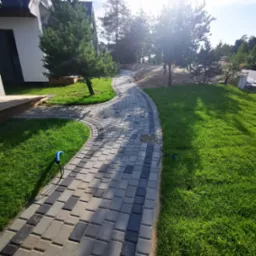 Zakrzywiony chodnik z szaro-czarnej kostki brukowej prowadzący przez zielony trawnik w kierunku domu i drzew, z widocznym niebieskim kablem przy trawniku.