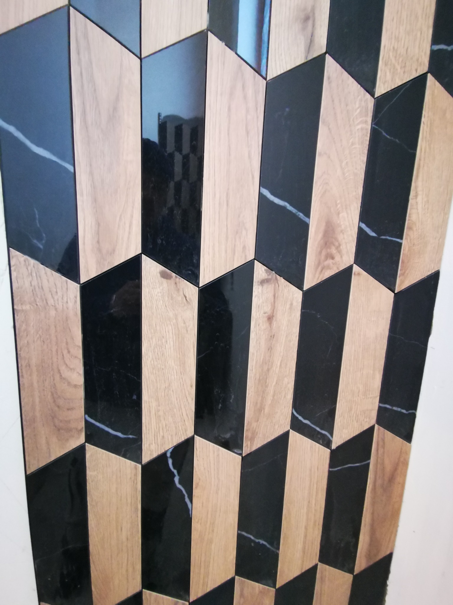 Ściana wyłożona naprzemiennie czarnymi płytkami imitującymi marmur z białymi żyłami i płytkami imitującymi drewno, ułożonymi w geometryczny wzór w kształcie strzałek.