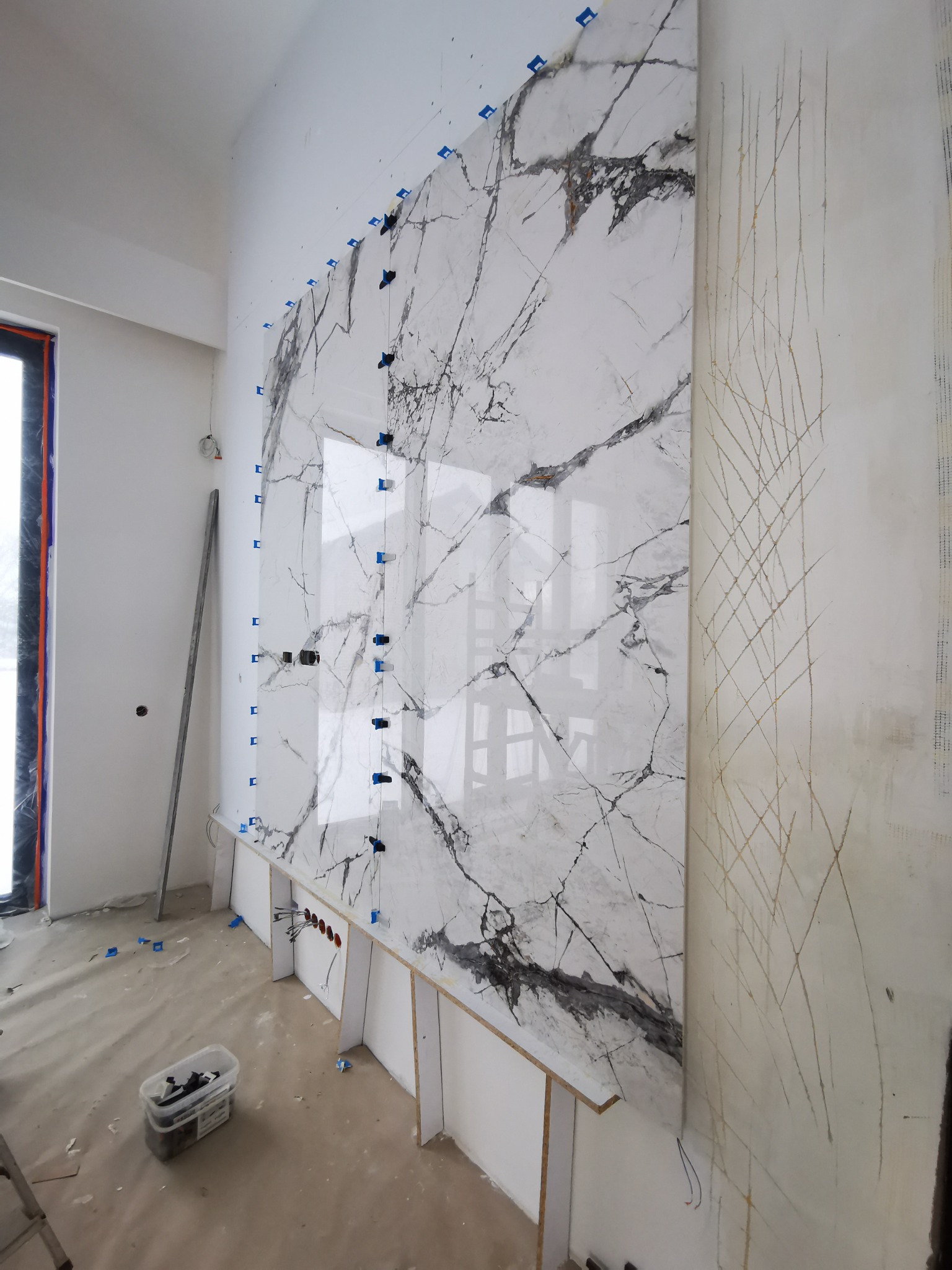 Wielkoformatowa płyta imitująca marmur, z niebieskimi klinami poziomującymi, montowana na ścianie z widocznymi śladami po ołówku i przygotowaną instalacją elektryczną.