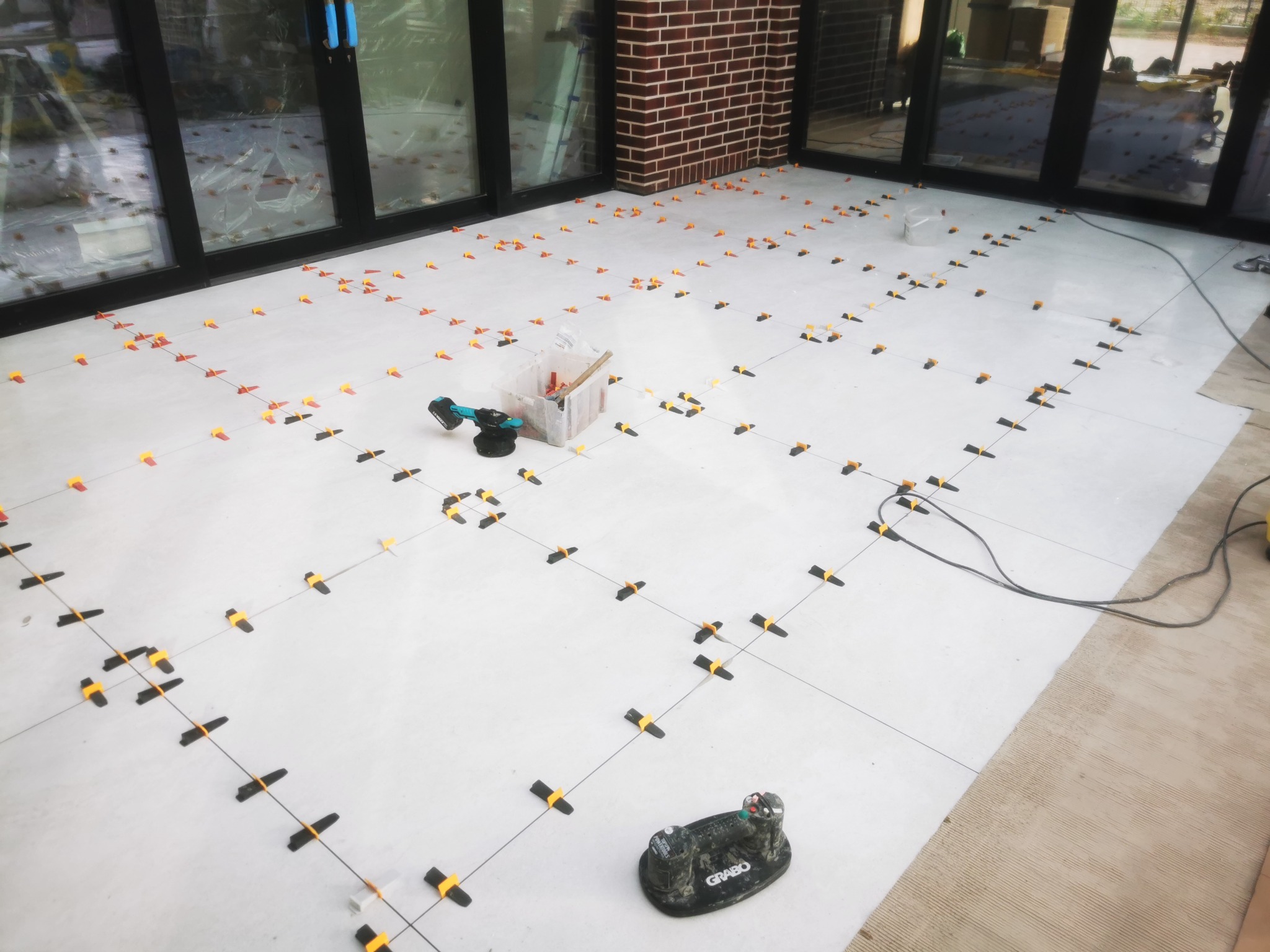 Układanie jasnych płytek podłogowych z użyciem systemu poziomowania, widoczne narzędzia: przyssawka Grabo, poziomica i pojemnik z klinami, tło stanowią przeszklone drzwi i fragment ściany z cegły.