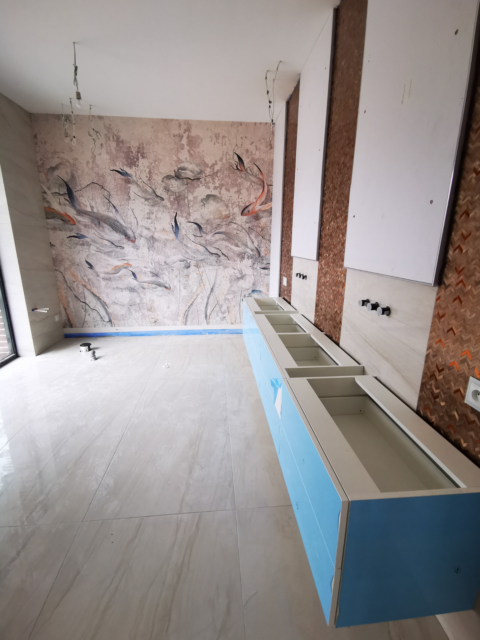 Łazienka w trakcie wykańczania: zamontowana szafka pod umywalkę z niebieskimi frontami, płytki imitujące marmur na podłodze i ścianach, dekoracyjna tapeta z motywem ryb na jednej ze ścian.