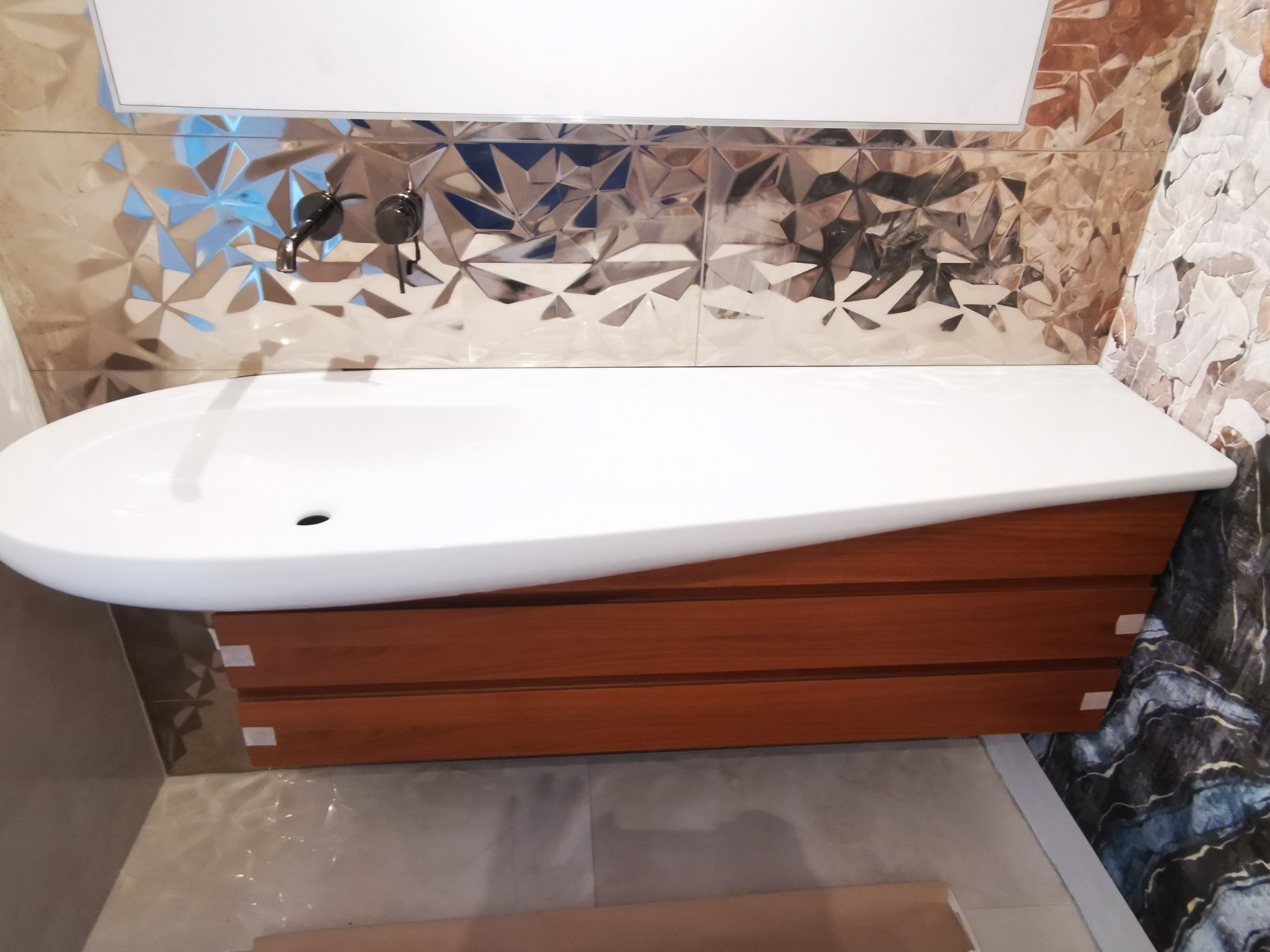 Nowoczesna umywalka nablatowa o asymetrycznym kształcie, zamontowana na szafce z lamelami w odcieniach brązu, na tle błyszczących, fasetowanych płytek i marmurowej okładziny ściennej, z widoczną...