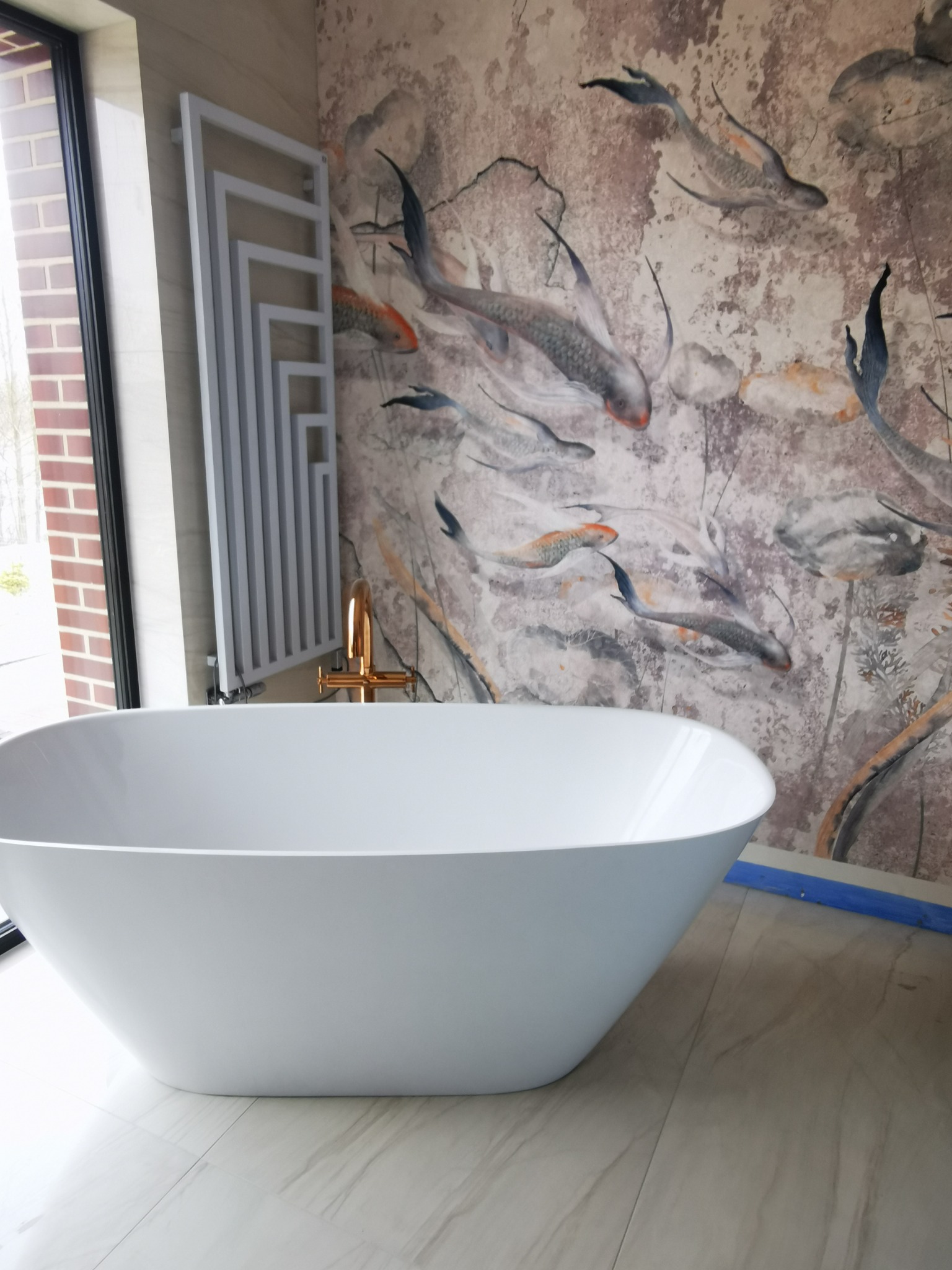 Wolnostojąca biała wanna owalna na podłodze z płytek imitujących marmur, na ścianie tapeta z motywem ryb koi, obok grzejnik drabinkowy i okno.