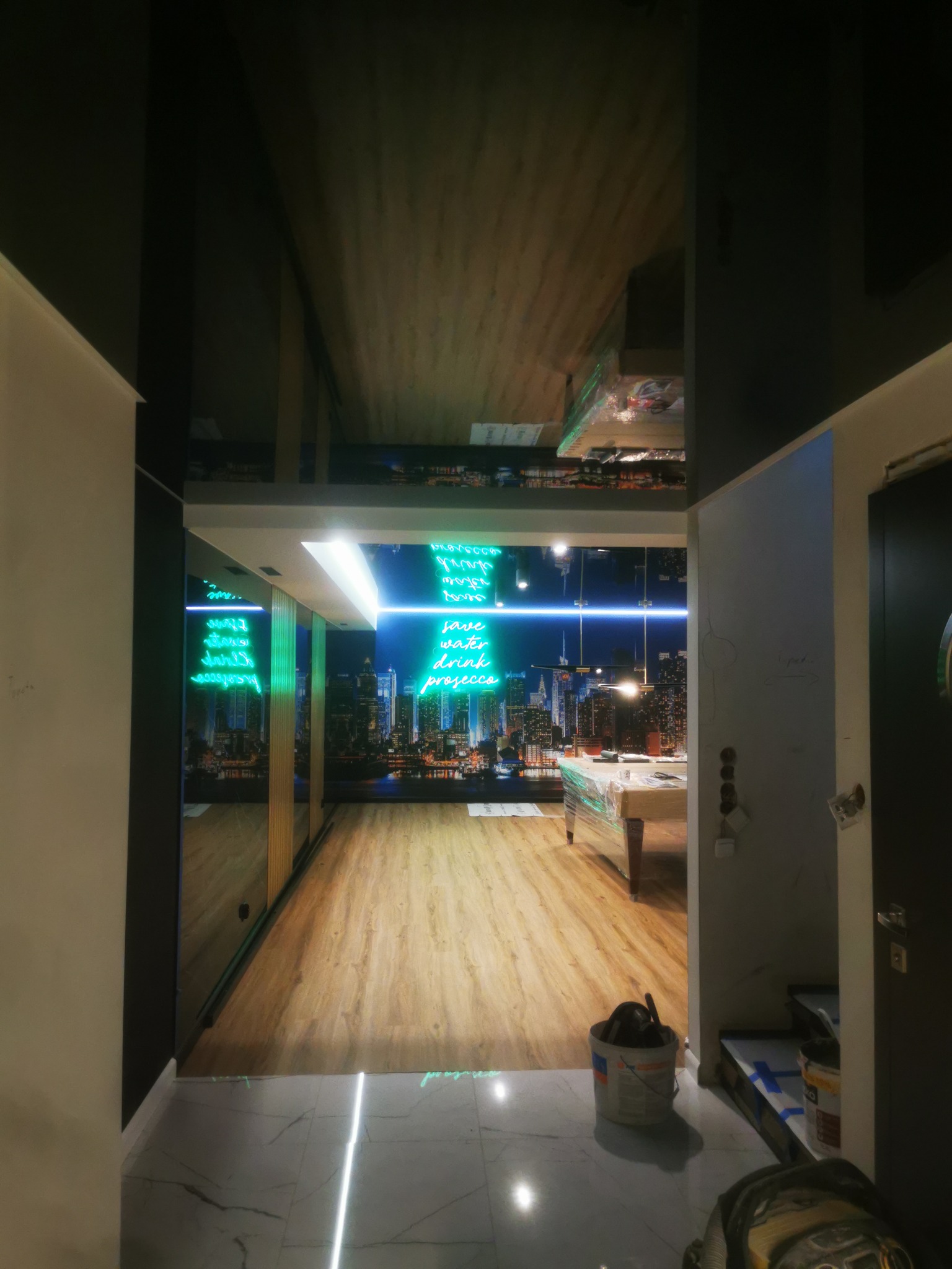 Wnętrze mieszkania w trakcie remontu, widoczna podłoga z paneli, ściana z tapetą przedstawiającą panoramę miasta, wiadro z farbą i narzędzia malarskie na marmurowej posadzce.