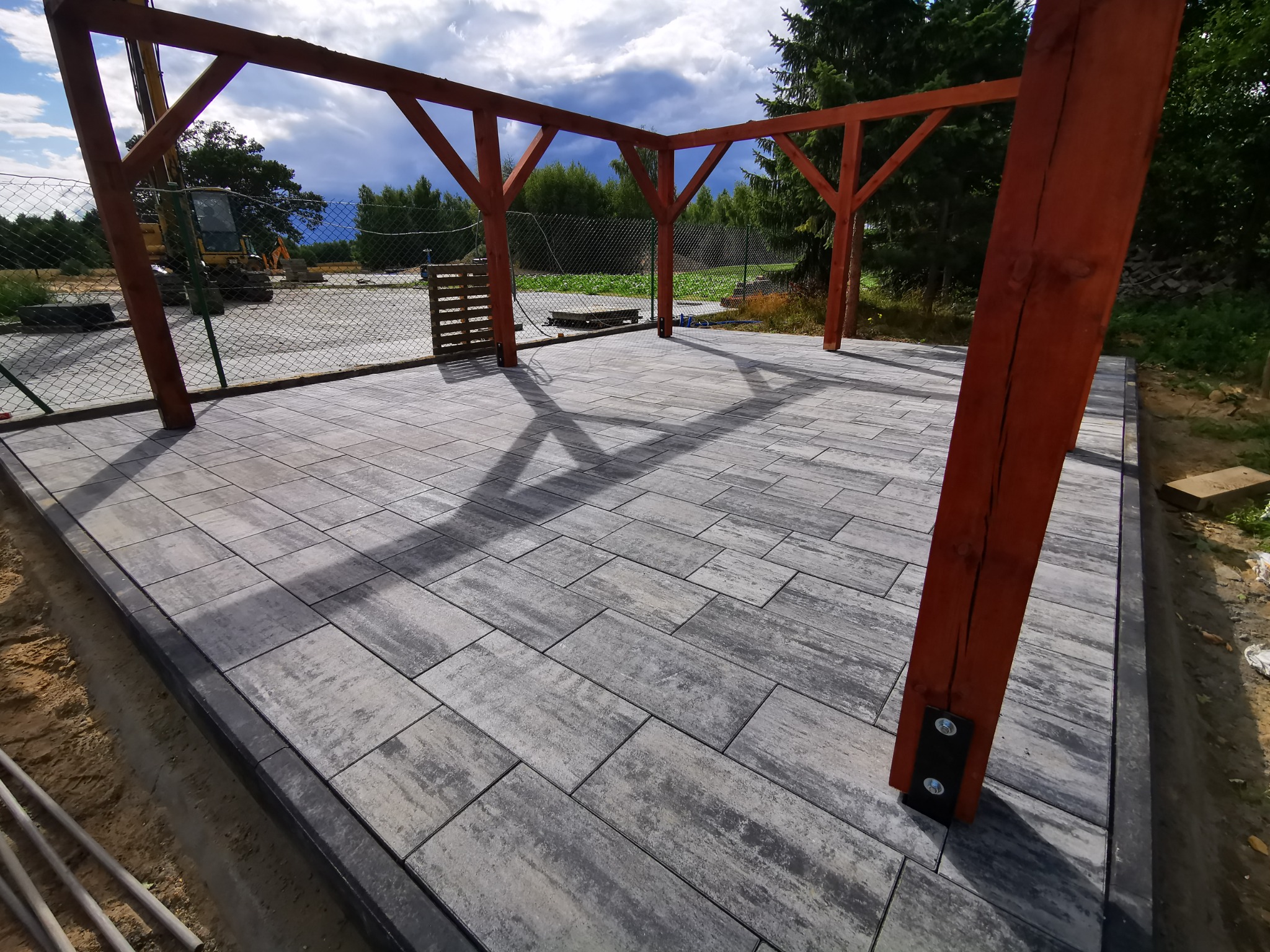 Ułożona szara kostka brukowa pod drewnianą konstrukcją altany, widoczne ślady świeżych prac ziemnych i koparka w tle.