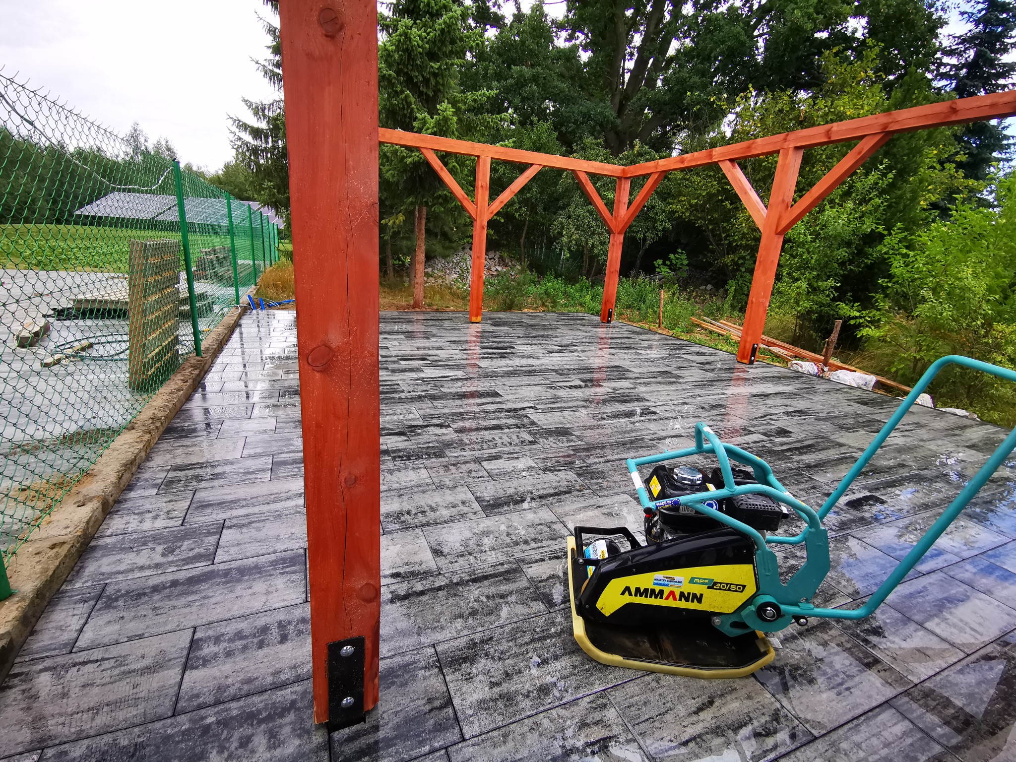 Świeżo ułożona, mokra kostka brukowa w odcieniach szarości, z widoczną ubijarką Ammann APS 20/50 oraz fragmentem drewnianej konstrukcji z pomarańczowych belek i ogrodzeniem z siatki.