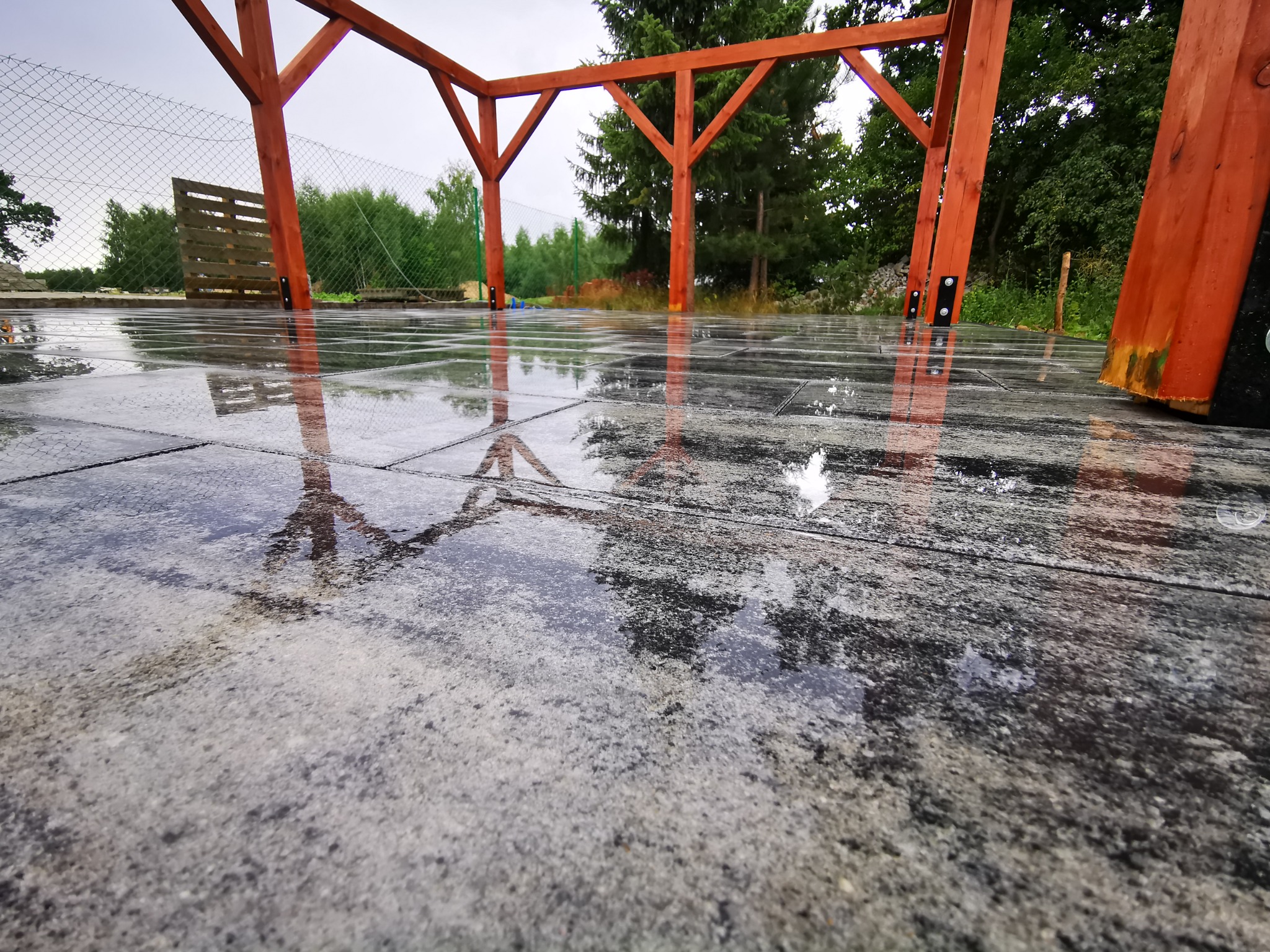 Świeżo ułożona, mokra, ciemnoszara podłoga z płytek na zewnątrz, z odbiciem drewnianej konstrukcji z czerwonego drewna w kałużach, widok z dołu.